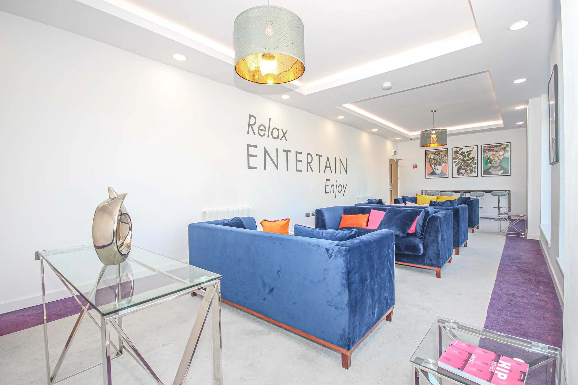 Modern lounge with blue velvet sofas and colorful pillows, white walls with motivational words, and framed artwork of floral arrangements.