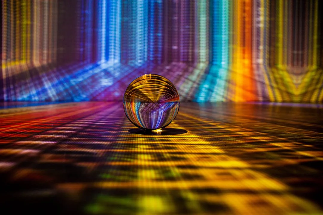A crystal ball resting on a surface with colorful light reflections and patterns projected on the background and floor.