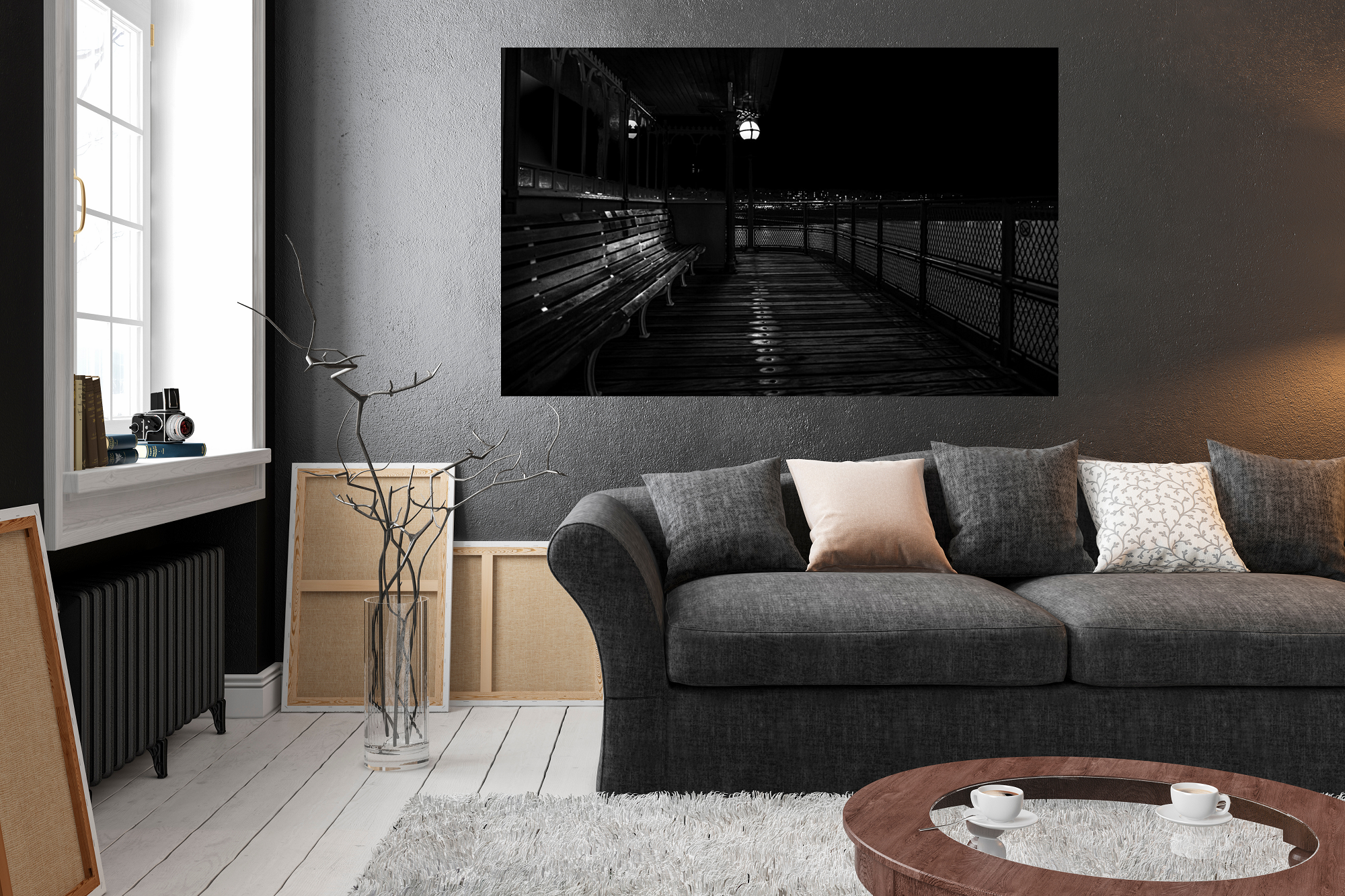 Living room with dark gray sofa and pillows, black and white framed photo on wall, window with books and camera on sill, empty picture frames on floor, white rug, and a round wooden coffee table with two cups of coffee.