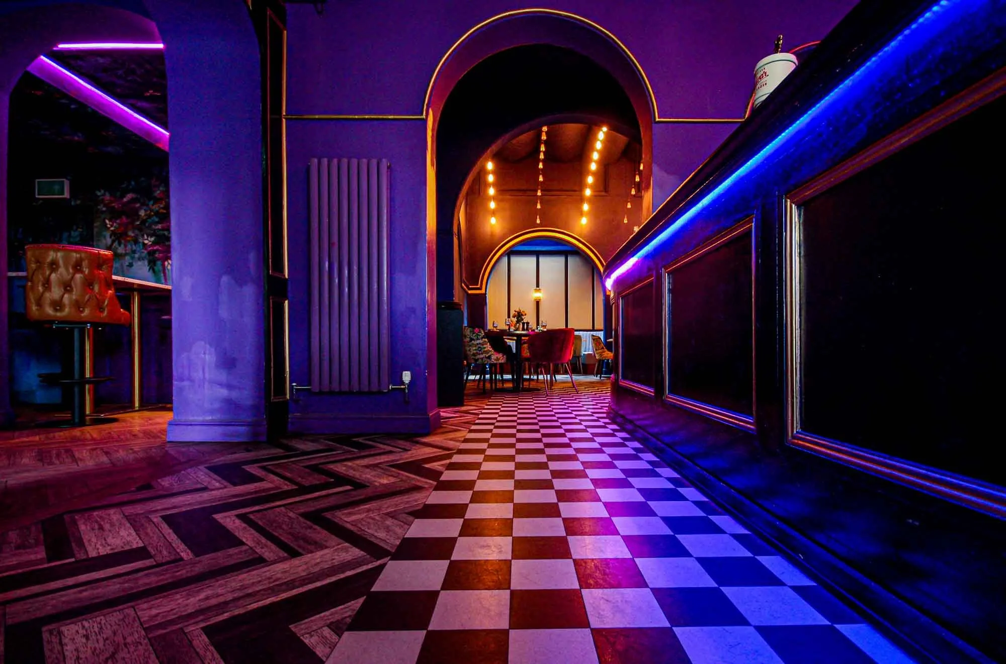 Interior of a bar or restaurant with purple walls, black countertops with purple and blue neon lighting, wood and checkered tile flooring, arched doorways, and a table with chairs in the background.