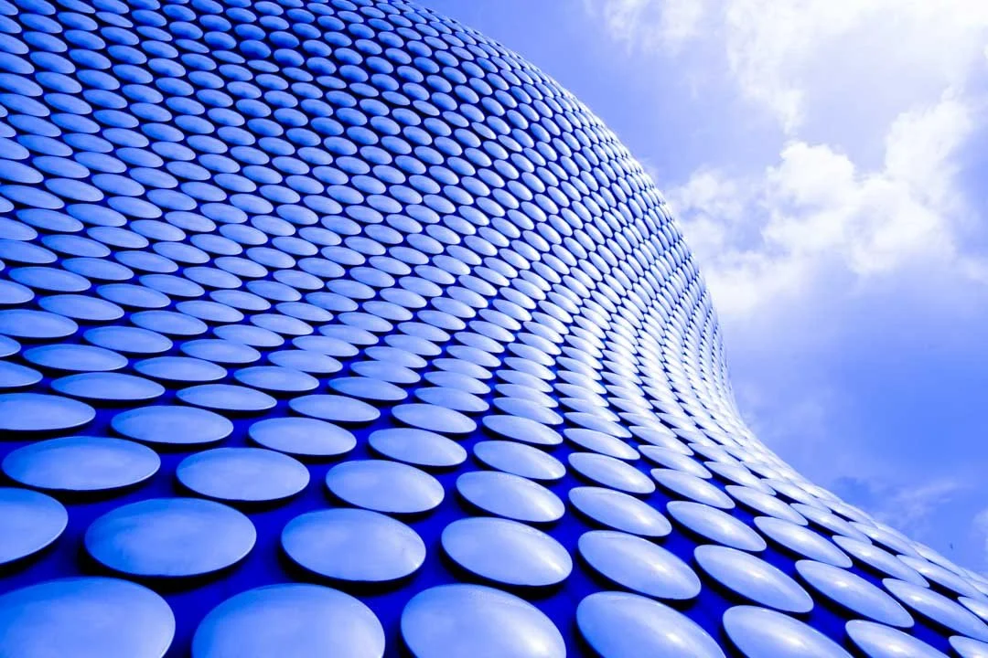 Close-up of a modern building with a curved facade covered in blue, oval-shaped panels, against a partly cloudy sky.