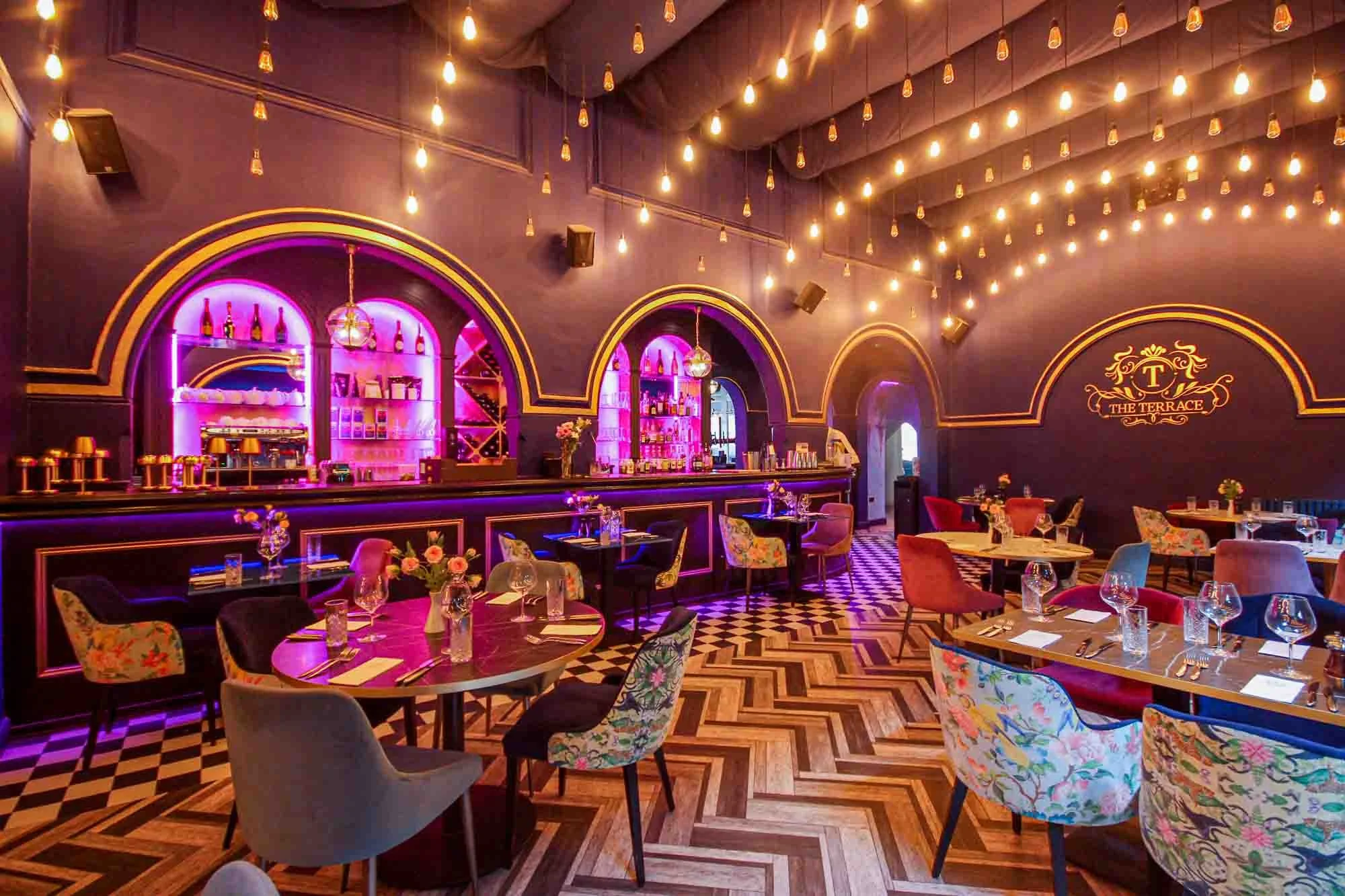 Stylish restaurant interior with purple walls, colorful flower-patterned chairs, and a checkered wooden floor. The ceiling has string lights, and there are tables set with glasses, cutlery, and napkins. The bar area features pink neon lighting and bo