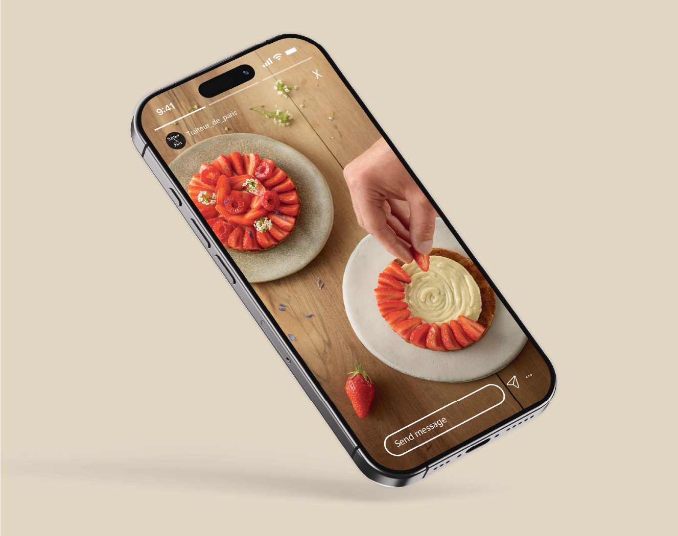 Un smartphone affichant une photo d'une tarte aux fraises garnie de fraises coupées en tuiles et de petites fleurs, avec une main ajoutant une fraise à la tarte.