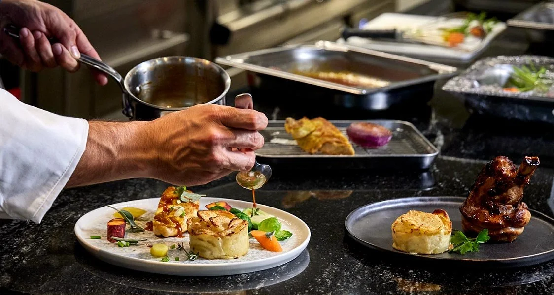 Un chef verse une sauce sur une assiette de plats gastronomiques contenant des légumes et des pommes de terre, avec un arrière-plan de comptoir de cuisine avec d'autres plats.