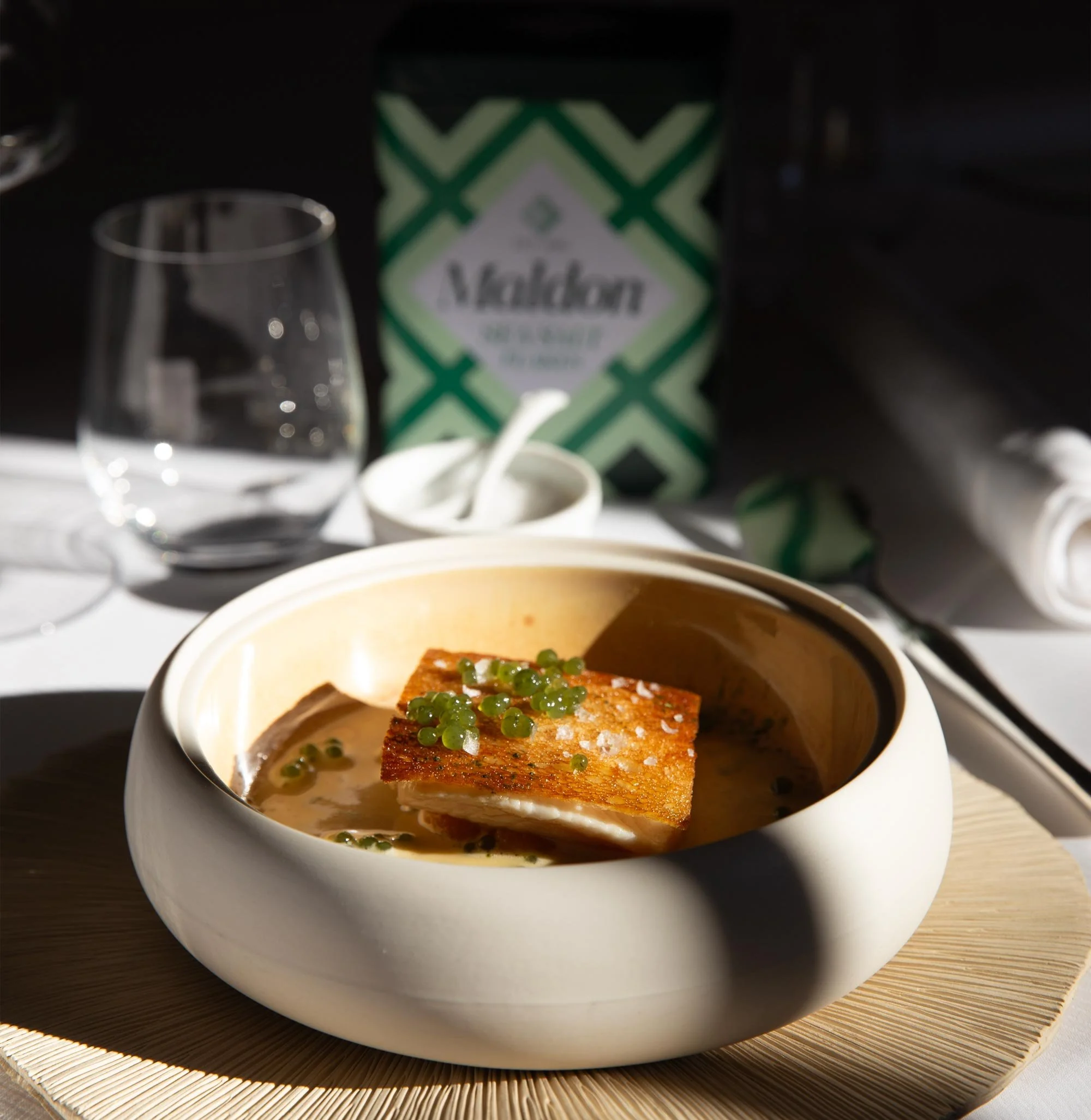 Plat de poisson avec une sauce, présenté dans un bol blanc rond sur une planche en bois, avec un fond flou d'ustensiles, verre d'eau et boîte de thé Matcha.