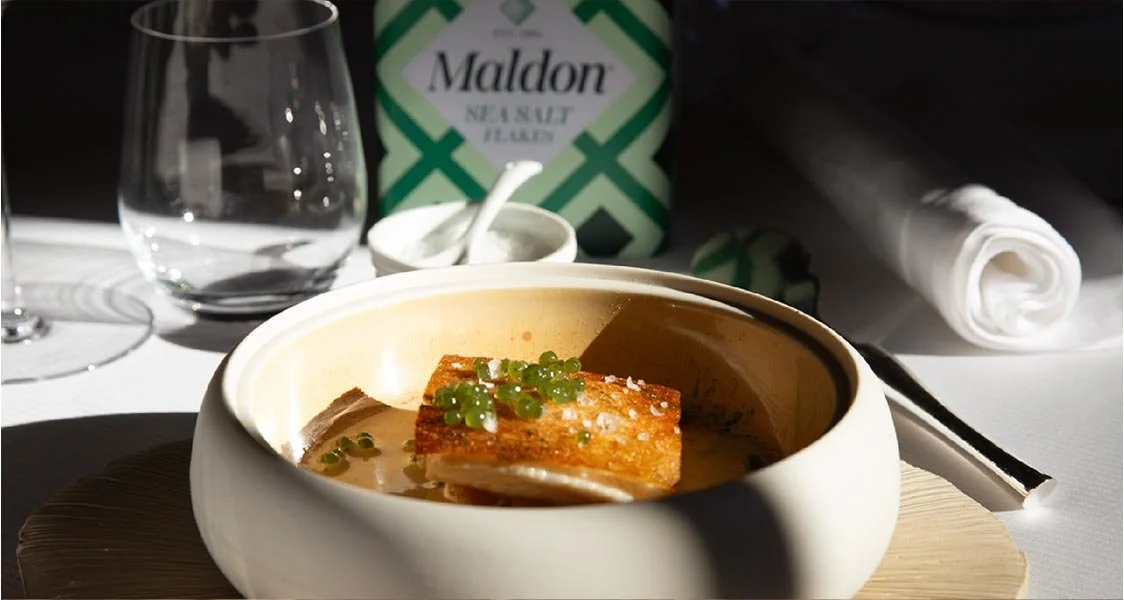 Plat gastronomique avec un morceau de poisson, garniture vert clair, dans un bol, avec verre d'eau, couvert, et article en papier à l'arrière.