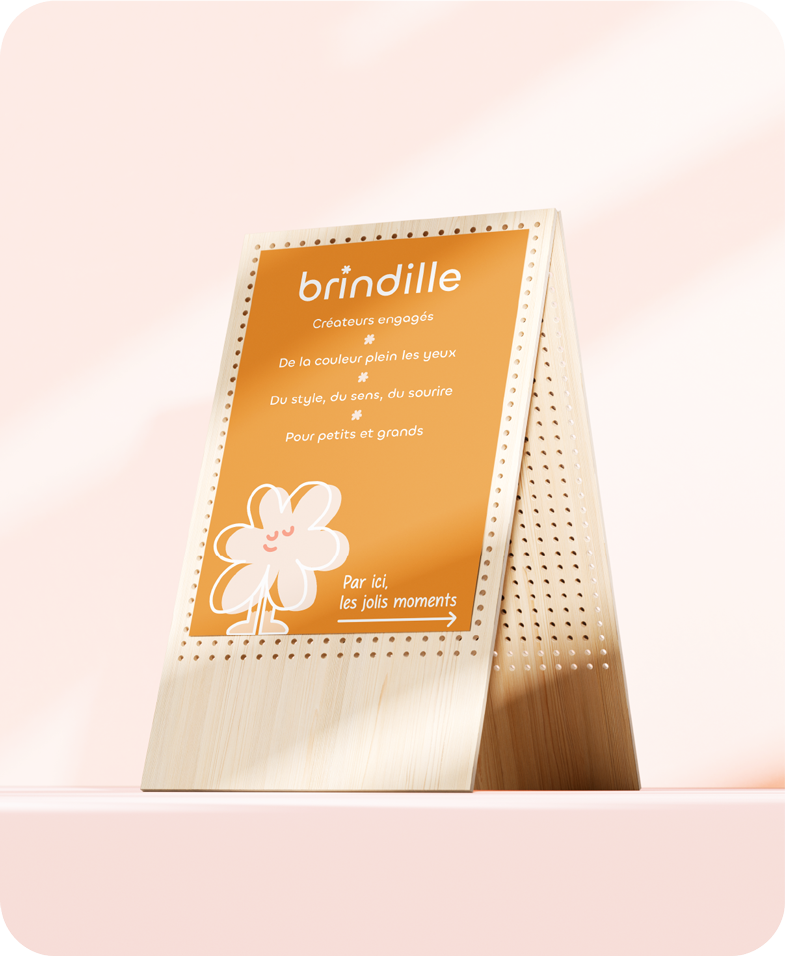 Panneau en bois clair avec un fond orange, présentant la marque 'brindille' et un dessin d'une fleur souriante, avec un texte promouvant les créations pour tous avec une flèche indiquant 'Par ici, les jolis moments'.
