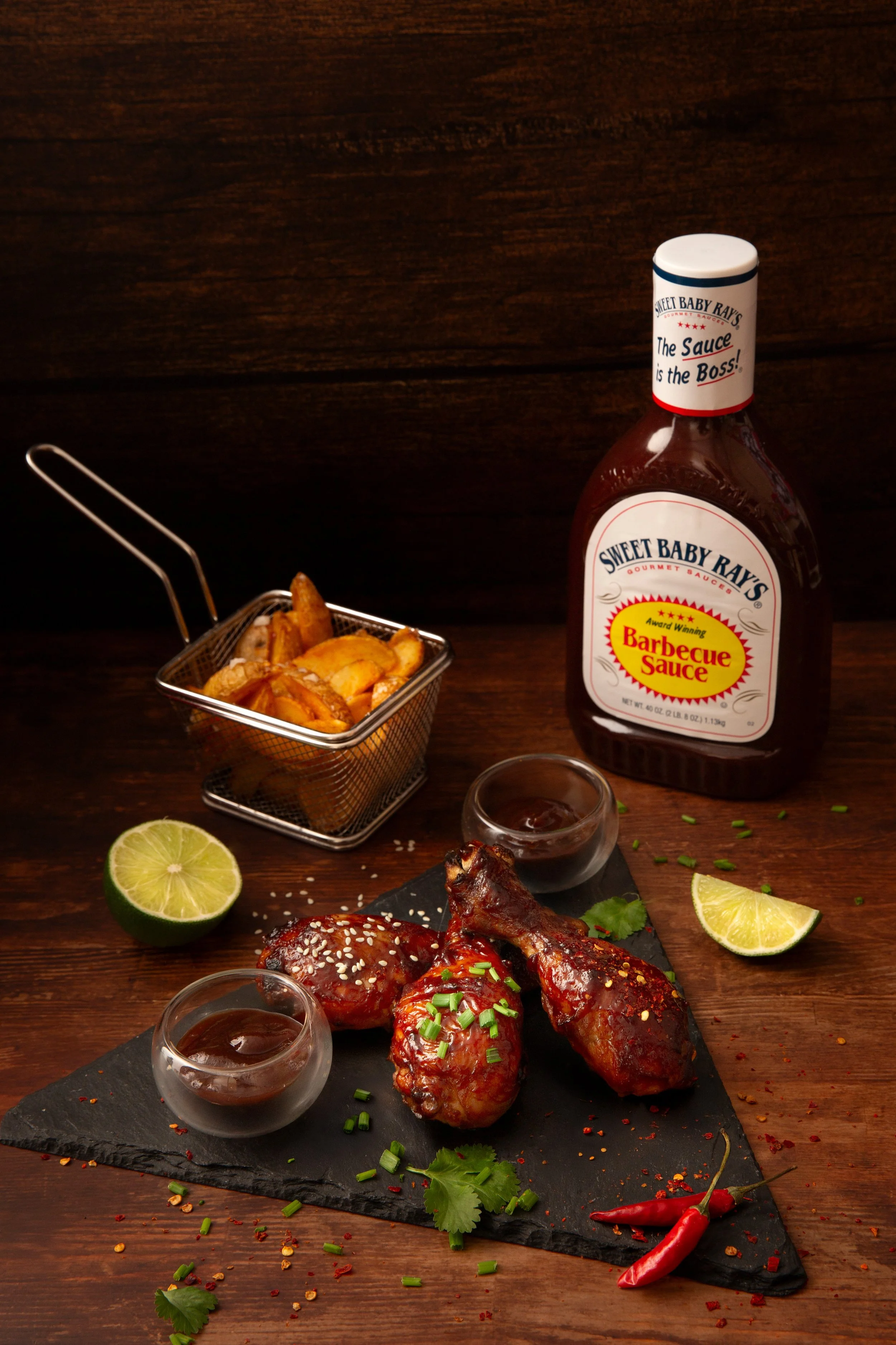 Shooting photo pour sauce Barbecue. Poulet frit, frites, sauce barbecue, quartiers de citron vert, corail rouge, petits bols de sauce barbecue, bouteille de sauce barbecue, herbes et épices sur une plaque en ardoise, fond en bois
