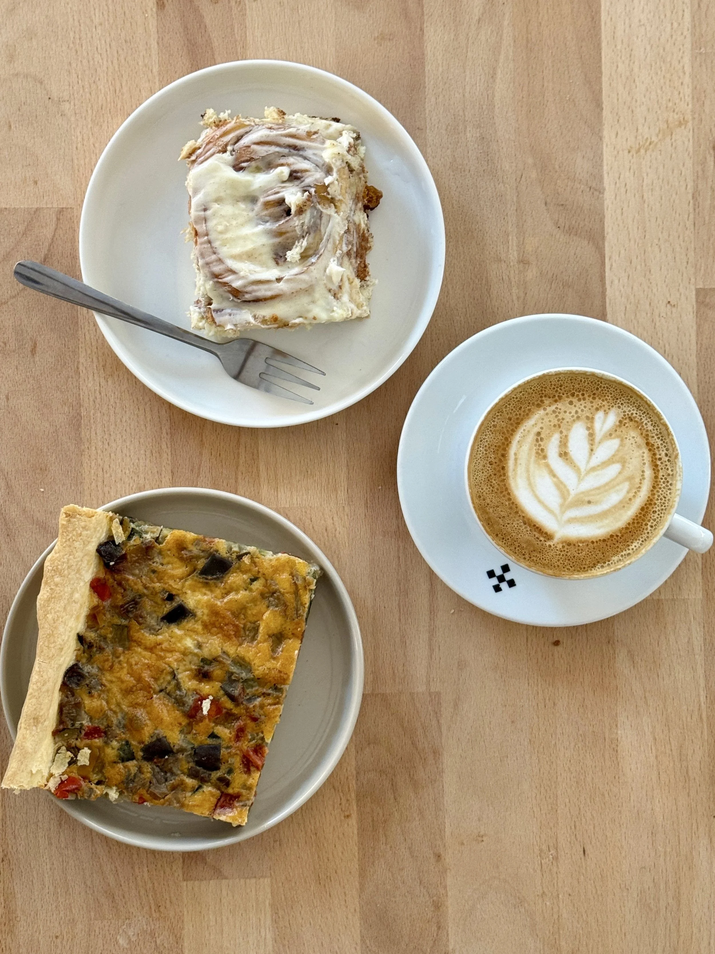 Une assiette avec une part de quiche aux légumes, une autre assiette avec un cinnamon roll avec sa crème cheese, une tasse de cappuccino avec latte art.