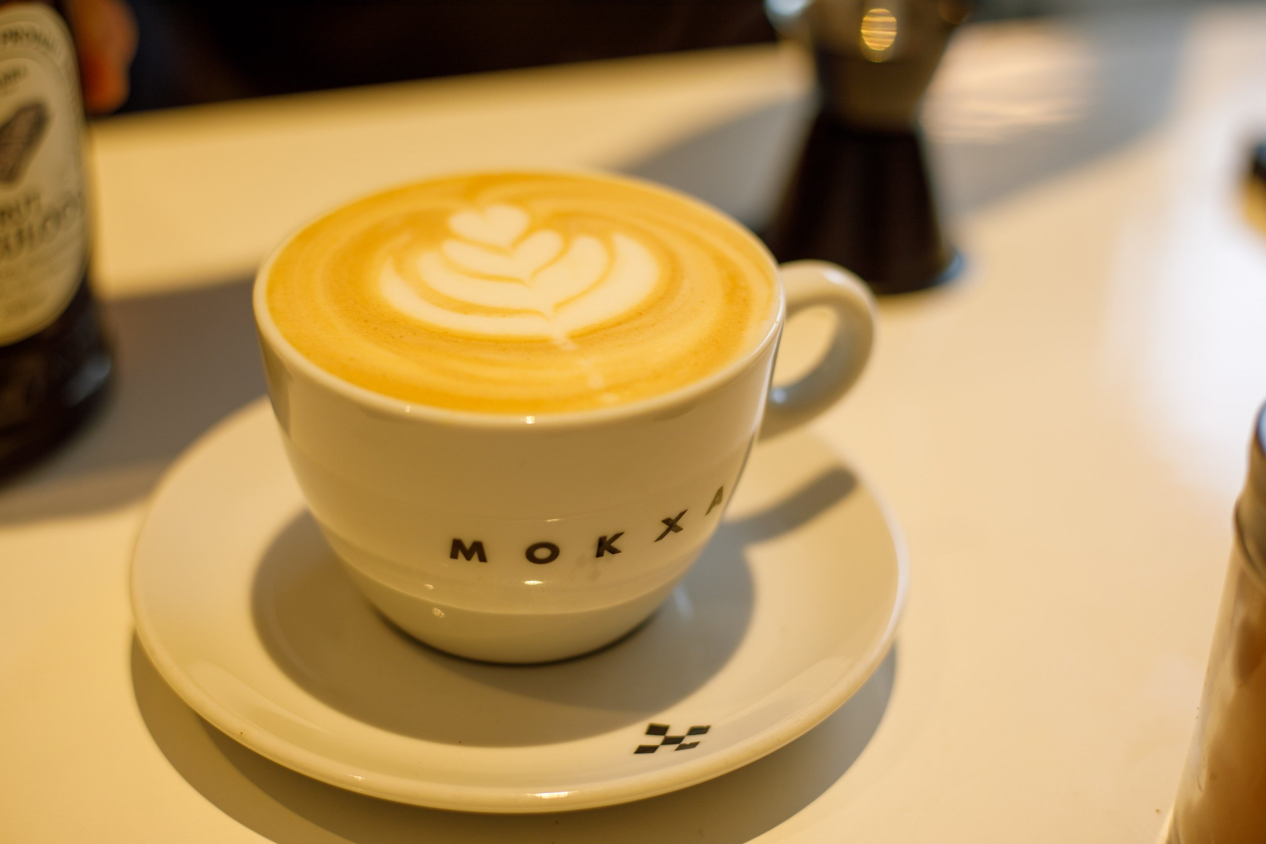 Une tasse de cappuccino avec un dessin en lait mousse sur le dessus, posée sur une soucoupe blanche, avec une variété d'objets flous en arrière-plan.
