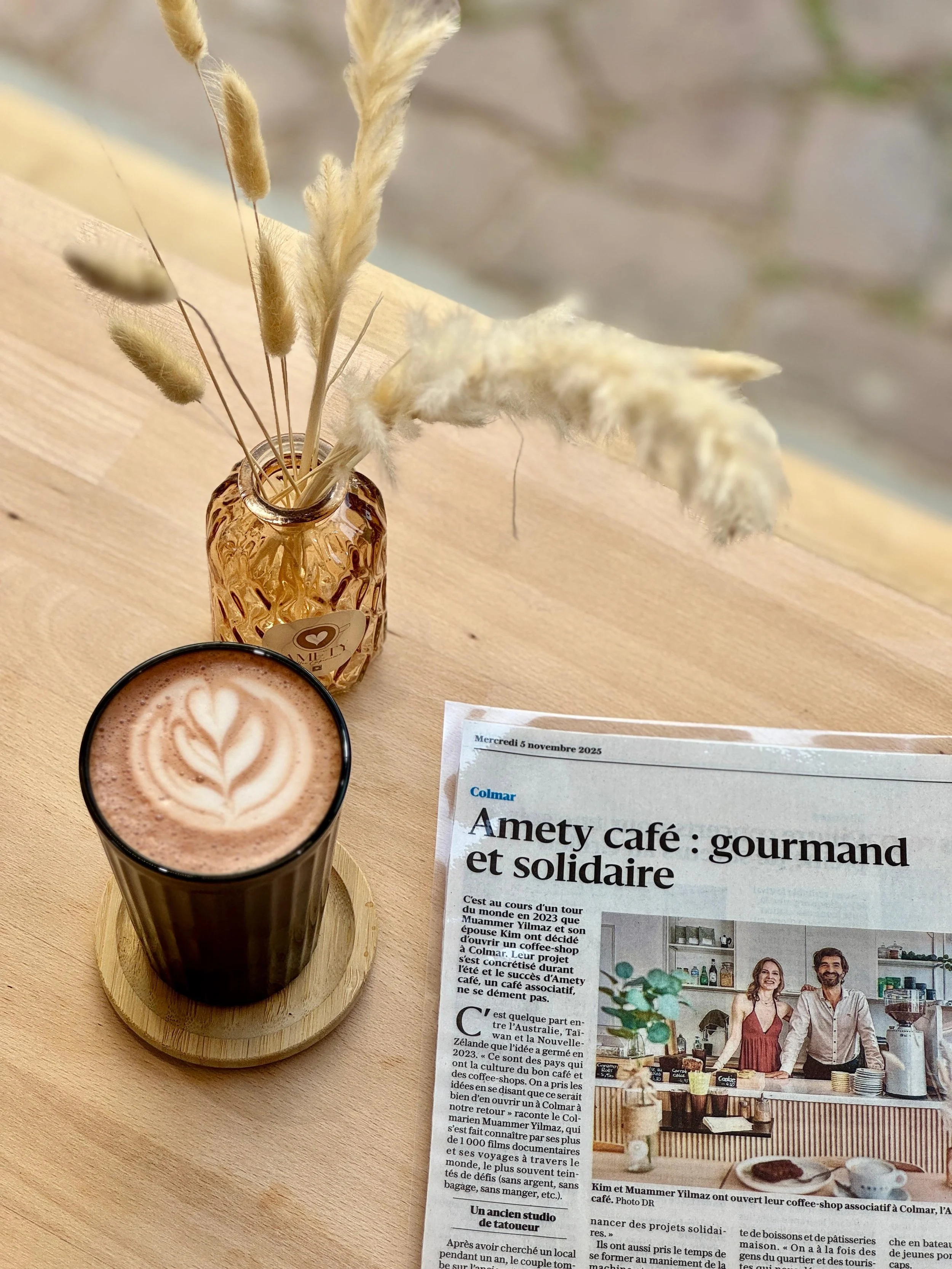 Un verre de chocolat chaud avec un motif en lait de type latte art, une petite vase avec des herbes séchées, et un article de journal en français sur notre café associatif à Colmar "Amety Café".