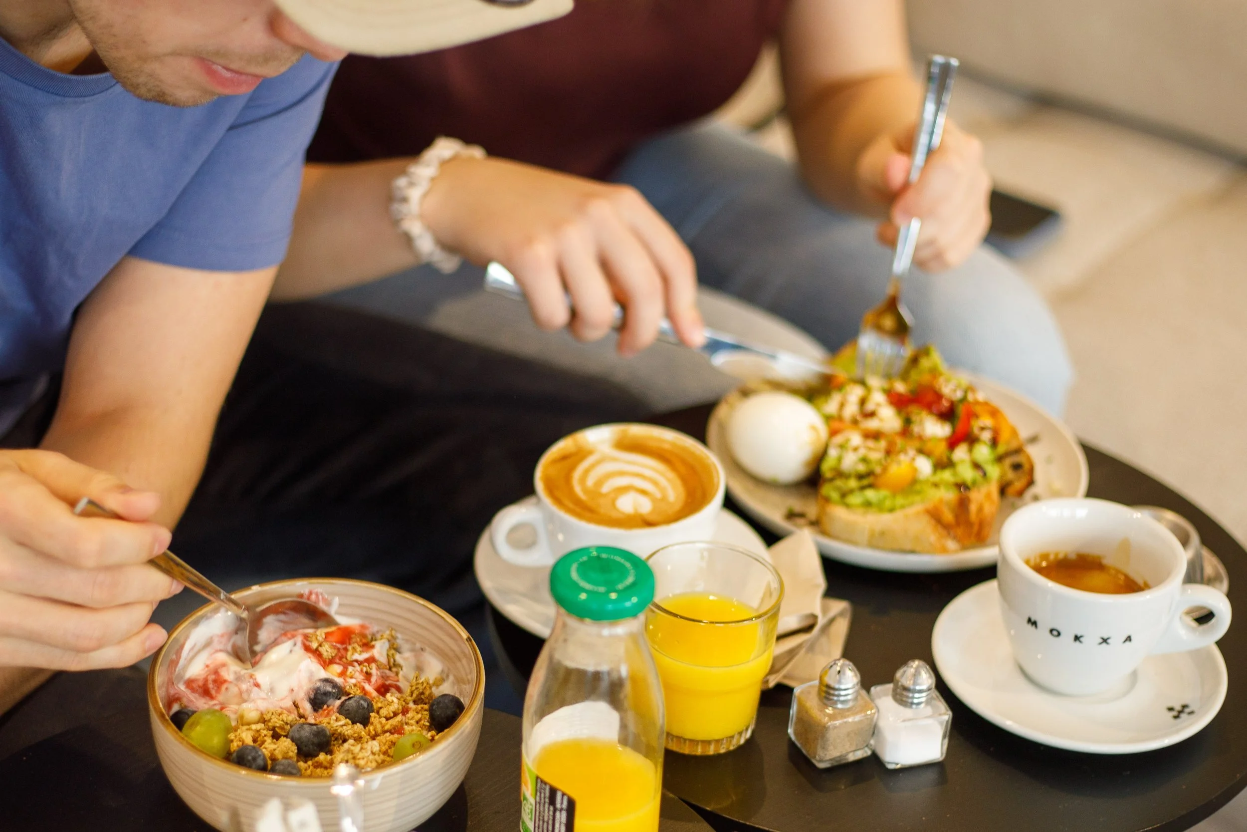 Deux personnes prennent un petit déjeuner brunch avec un avocado toast, œuf, américano, cappuccino, jus d'orange, et un bol de yogourt avec des fruits (raisins et myrtille), un coulis de framboise et du granola.