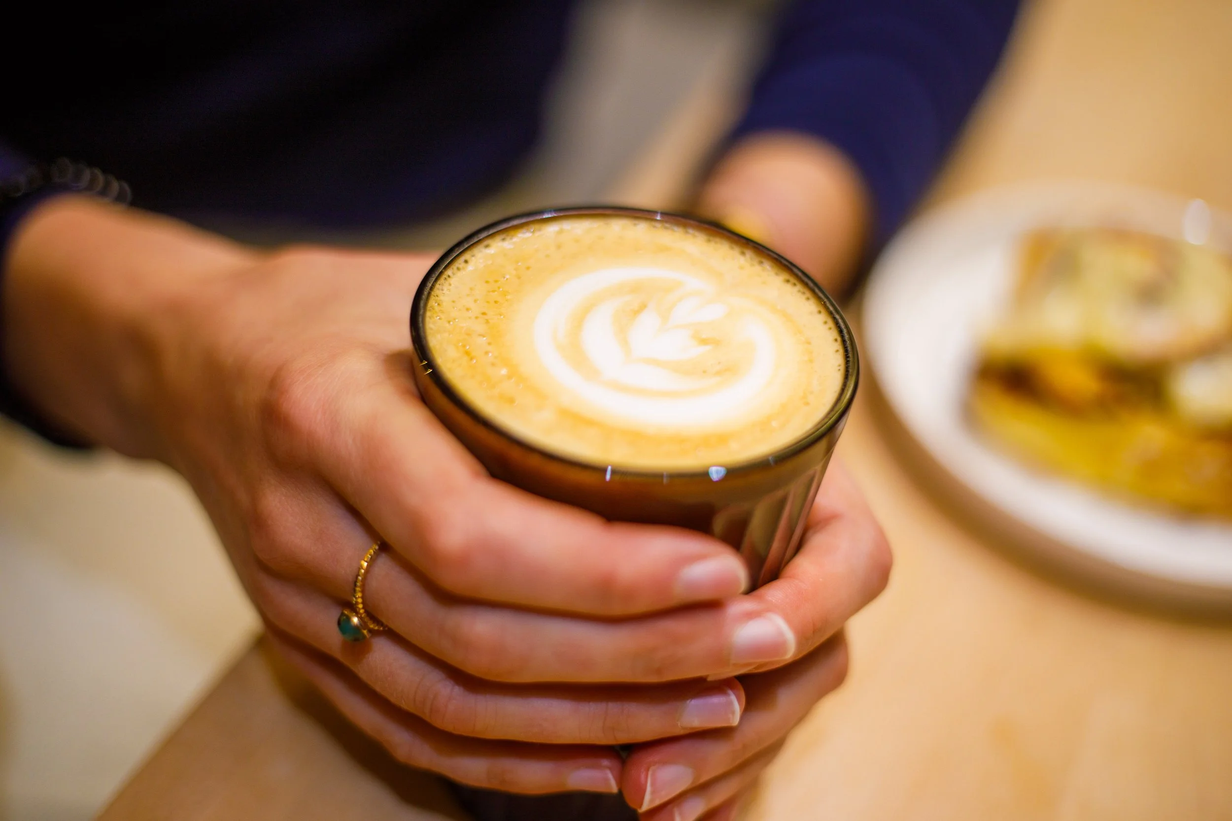 Main tenant un verre de café latte avec un dessin en latte art, une autre tasse et une assiette avec des biscuits en arrière-plan.