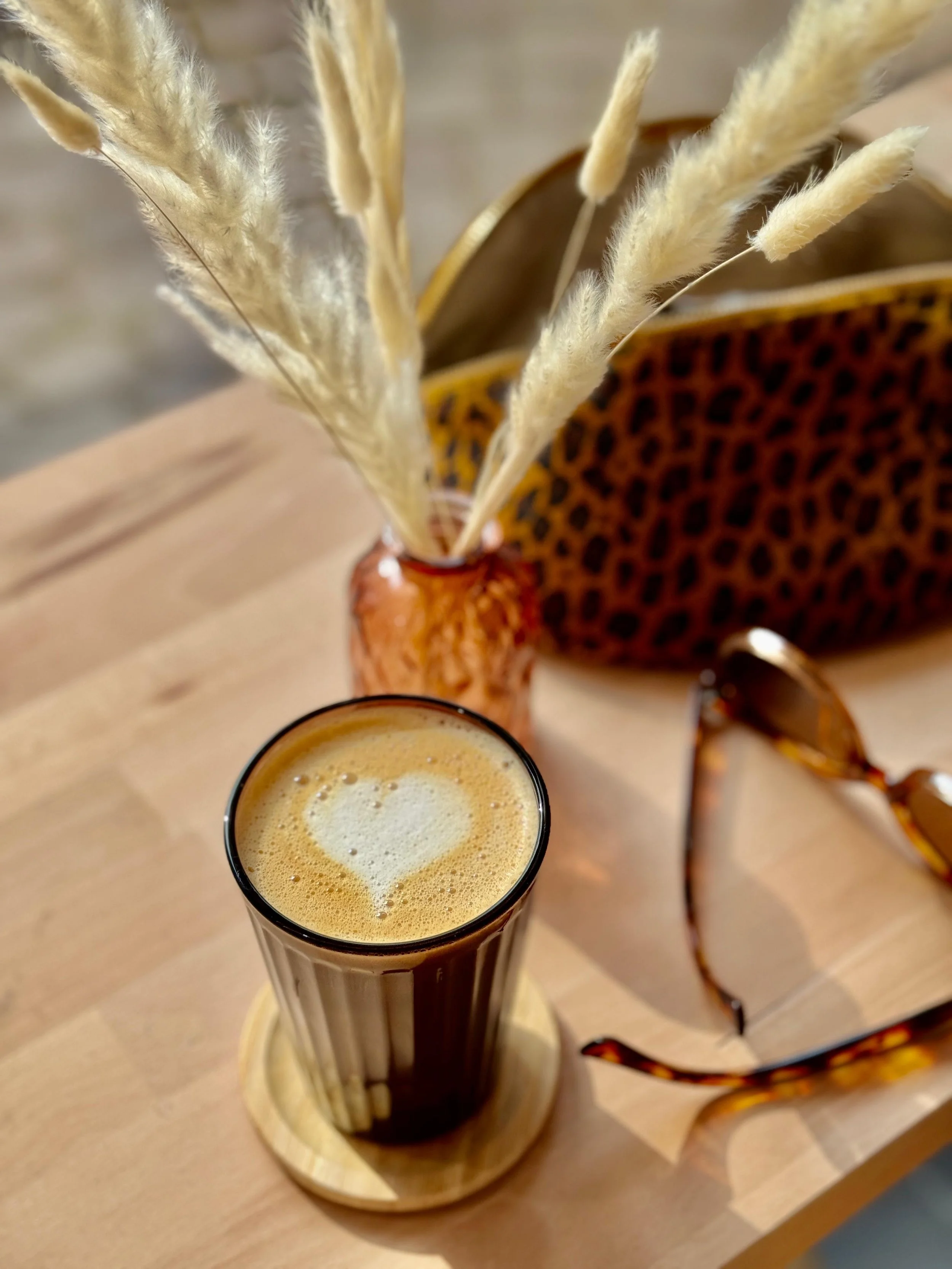 Un verre de café latte avec un cœur en mousse, placé sur une table en bois avec des lunettes de soleil et un vase contenant des herbes de la pampa et un sac en léopard.