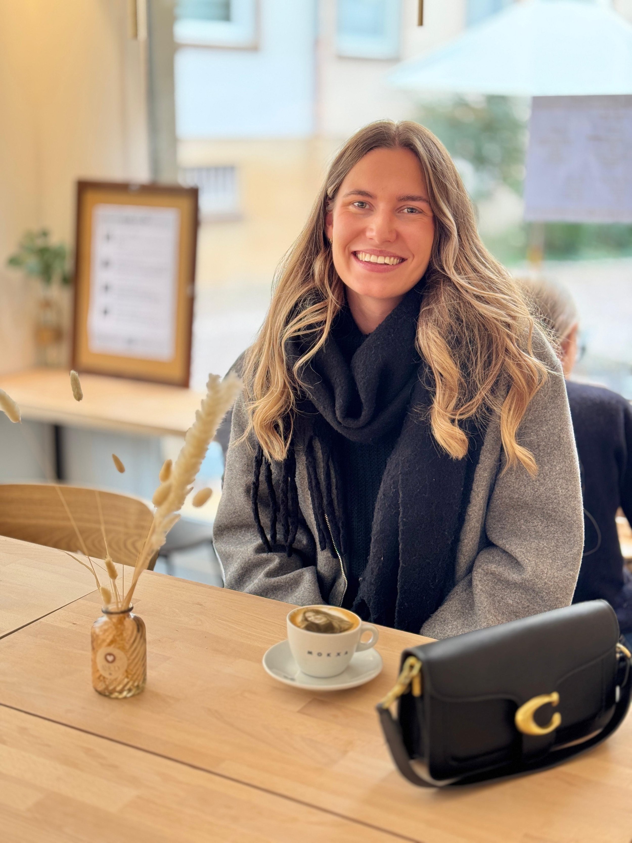 Jeune femme souriante assise à une table dans le café Amety Café, avec une tasse de cappuccino avec sa photo imprimée dessus devant elle, un sac à main noir et un vase avec des herbes décoratives.