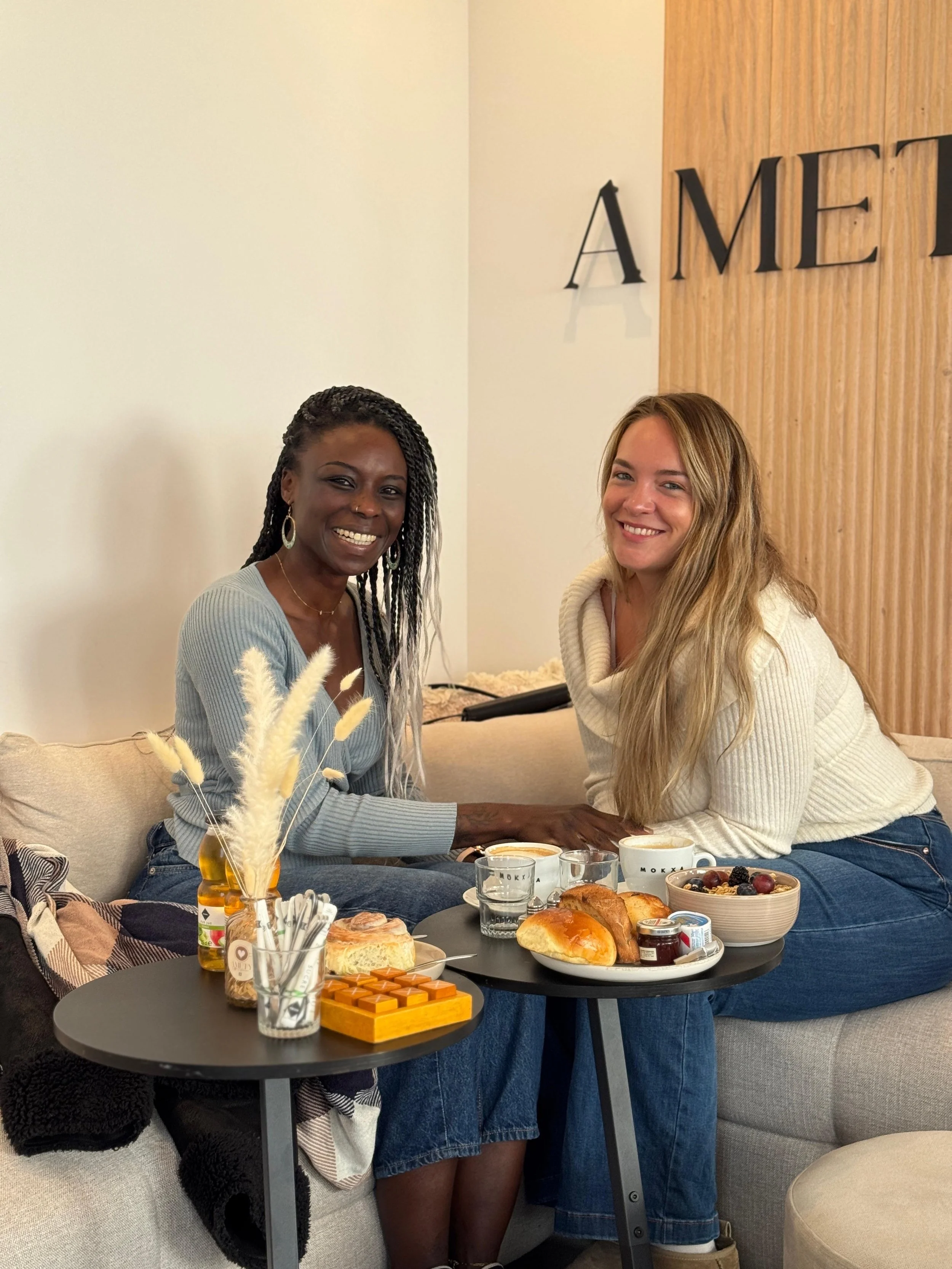 Deux femmes assises sur un canapé autour d'une table avec petit déjeuner, souriantes, dans un café ou salon.