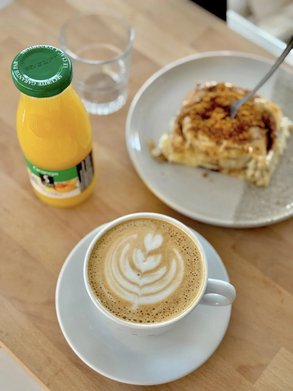 Un cappuccino avec un dessin en latte art dans une tasse blanche, un verre d'eau, une bouteille de jus d'orange, et une part de cinnamon roll au spéculoos avec une crème spéculoos sur le dessus sur une assiette.