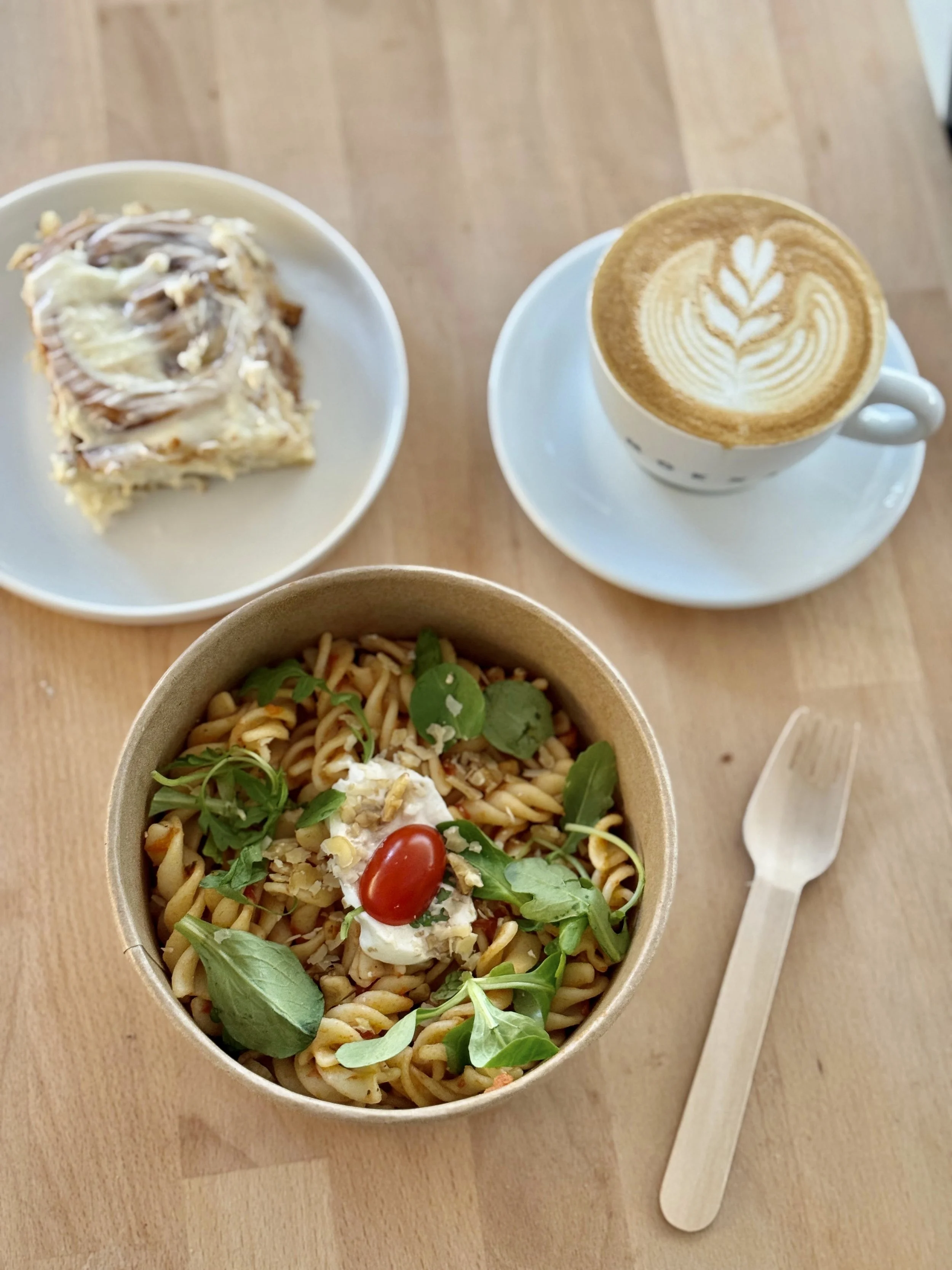 Un bol de salade de pâtes avec des tomates cerises, du fromage, de la sauce pesto et de la salade, un cinnamon roll avec un glaçage, un cappuccino avec du latte art sur la table en bois.