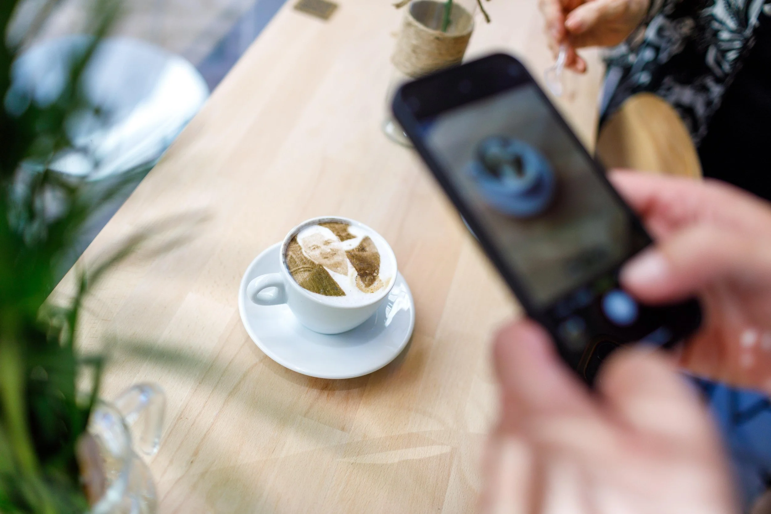 Une personne prend une photo d'une tasse de cappuccino avec l'impression avec de la poudre de café sur la mousse, du portrait du maire de Colmar Éric Straumann, sur une table en bois.