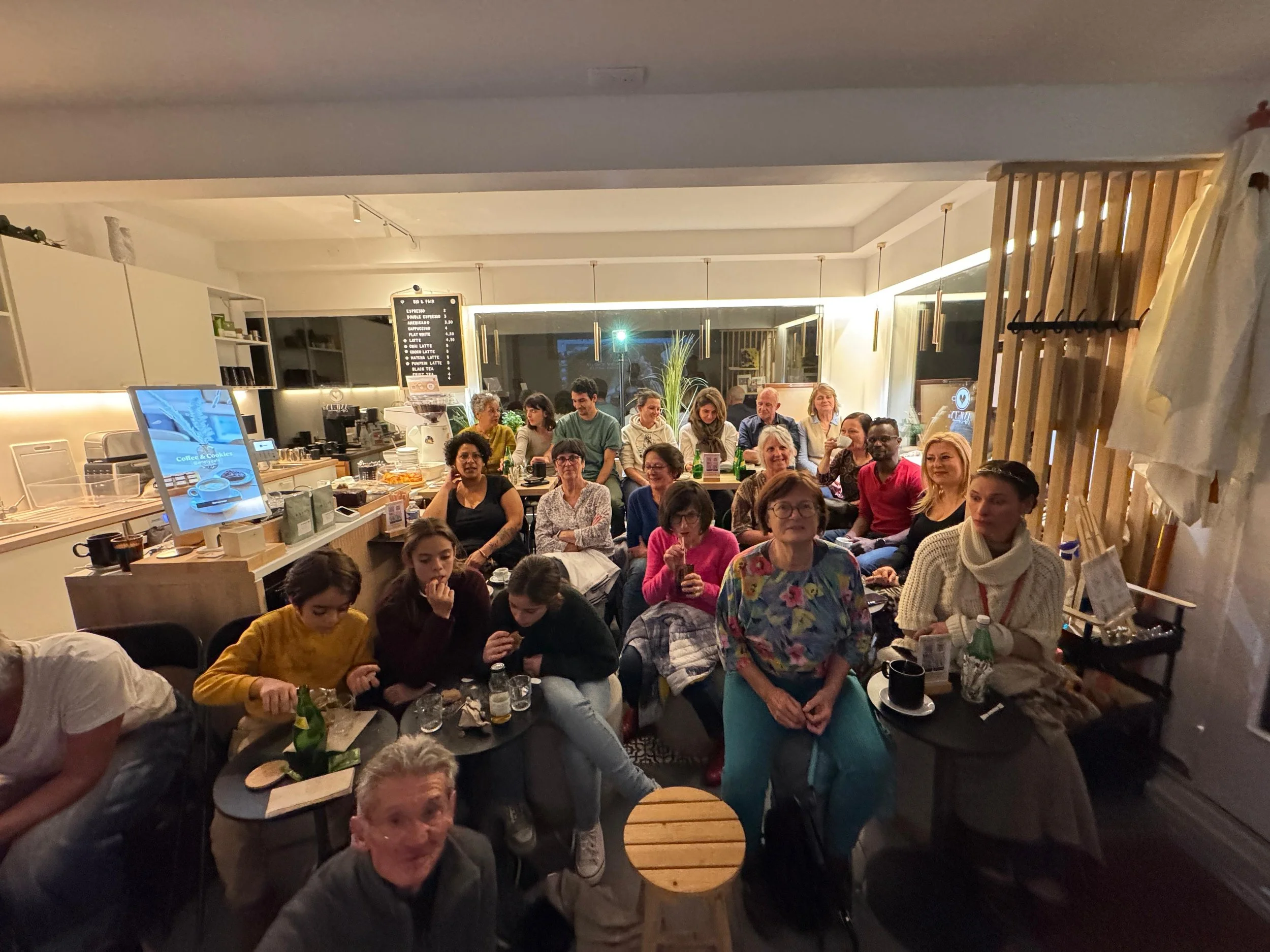 Groupe de personnes assises dans un café ou un restaurant, souriant et regardant vers l'appareil photo, avec une ambiance chaleureuse et conviviale.