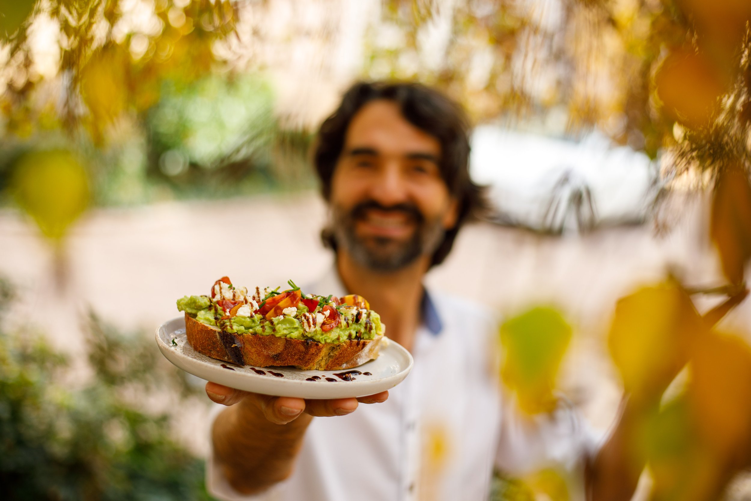 Un homme Muammer YILMAZ souriant tient une assiette avec une tartine d'avocat garnie de tomates, de feta et de sauce balsamique, pour lui, dans un cadre extérieur flou avec des feuilles d'automne.