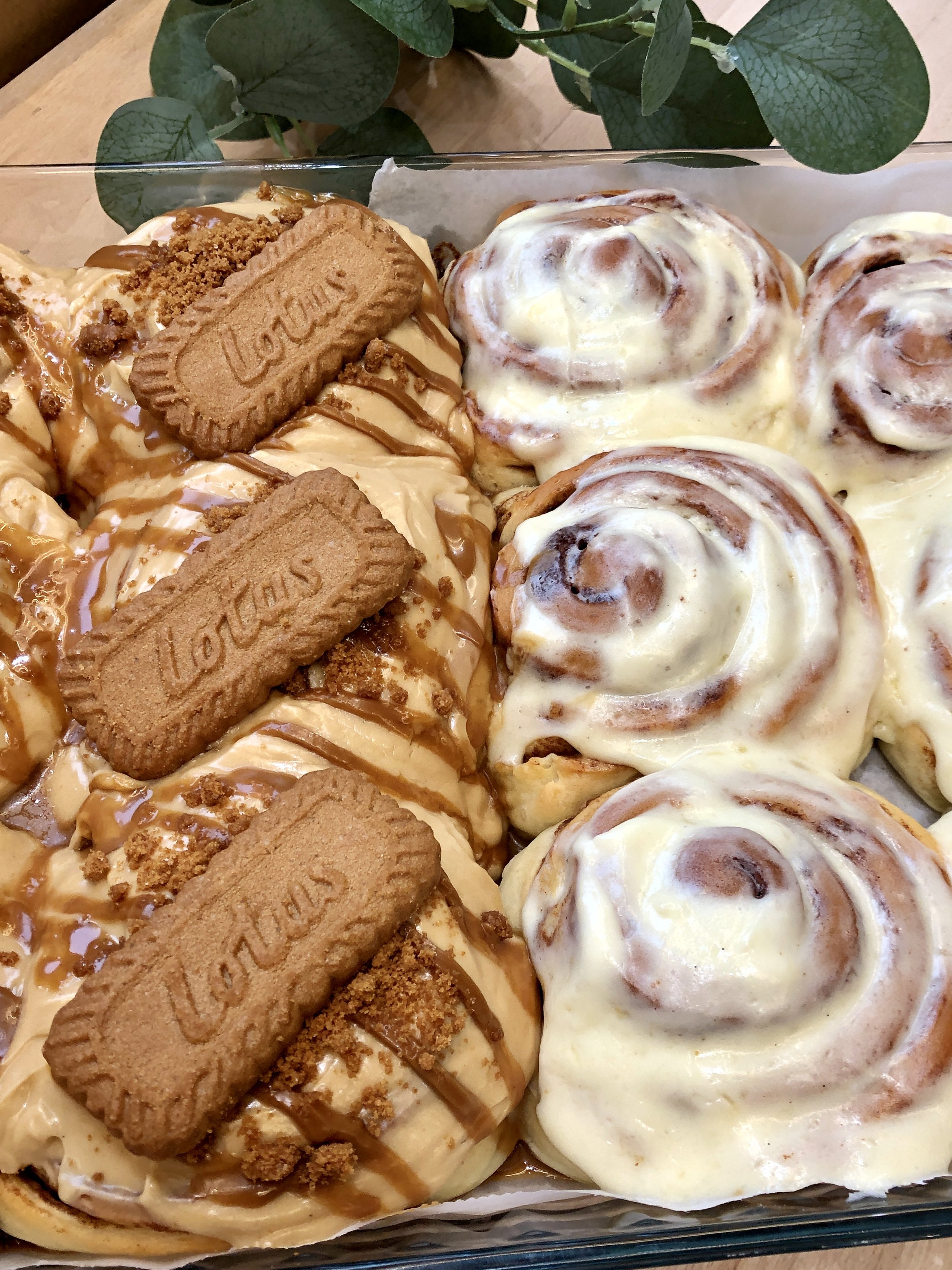 Gros plan de deux types de cinnamon rolls dans un plat en verre, un garnis de glaçage et l'autre avec des biscuits spéculoos Lotus et une crème spéculoos sur le dessus.