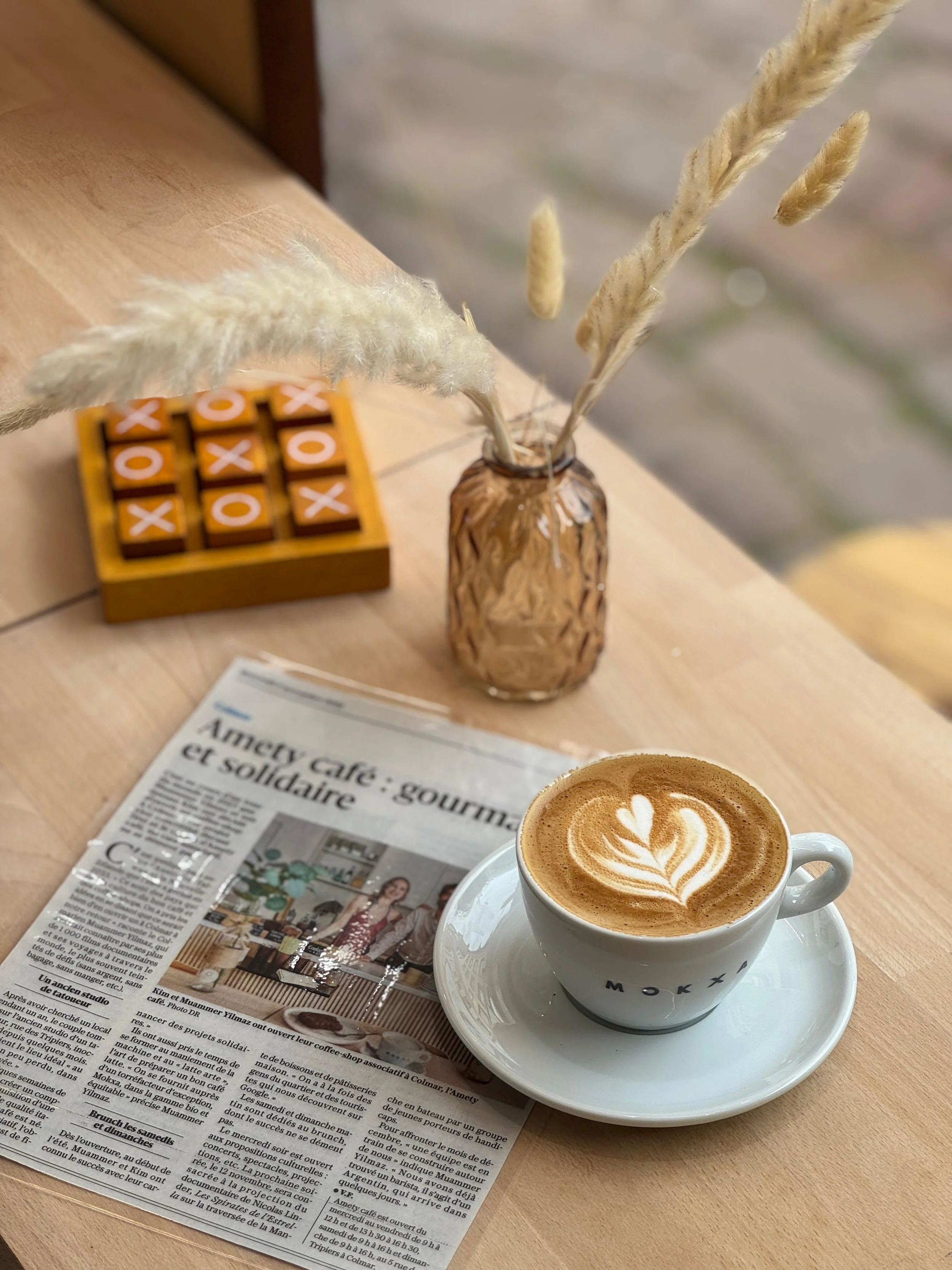 Une tasse de café latte avec un motif en forme de cœur, posée sur une table en bois, à côté d'un journal avec l'article du 5 novembre 2025 concernant le café "Amety Café", il y a aussi un petit vase avec des branches de pampa et un jeu de Morpion.
