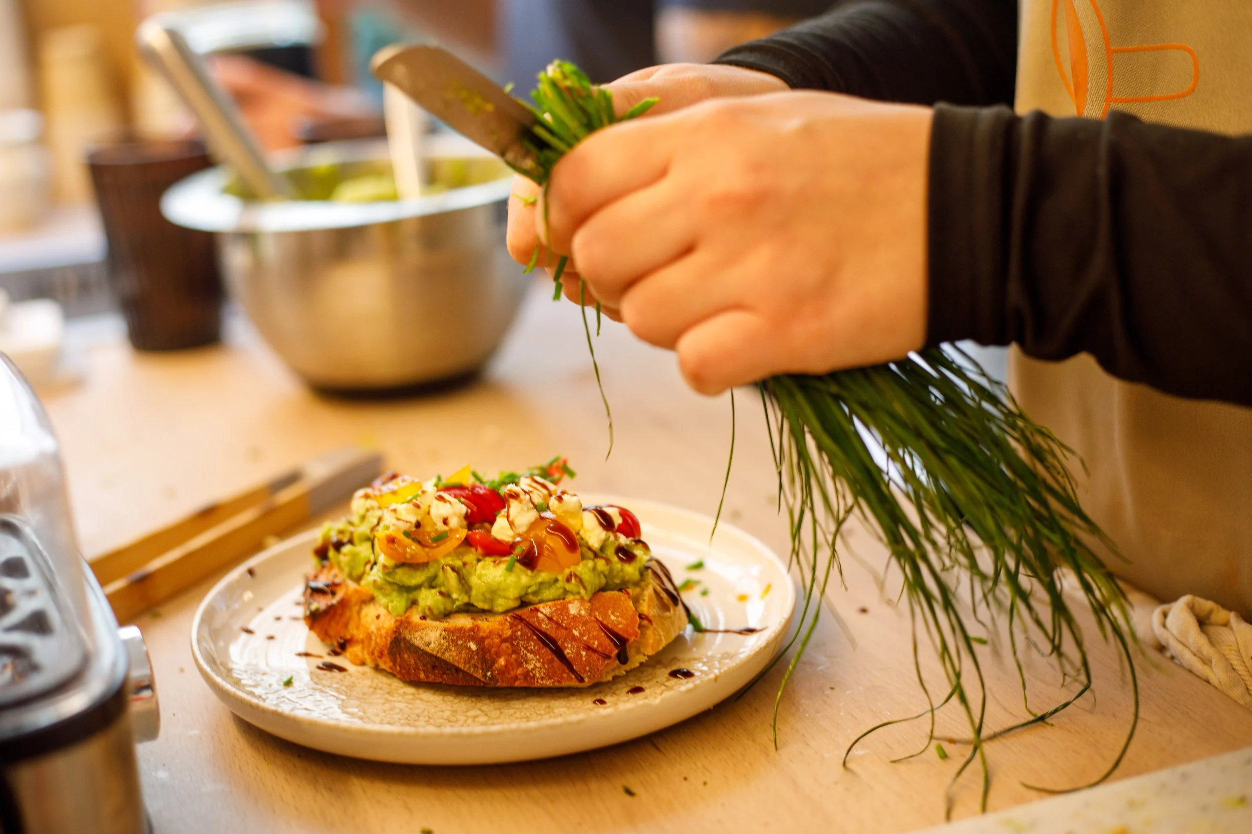 Une personne orne une assiette de tartine avec de l'avocat, des tomates, de la vinaigrette balsamique et des herbes fraîches.