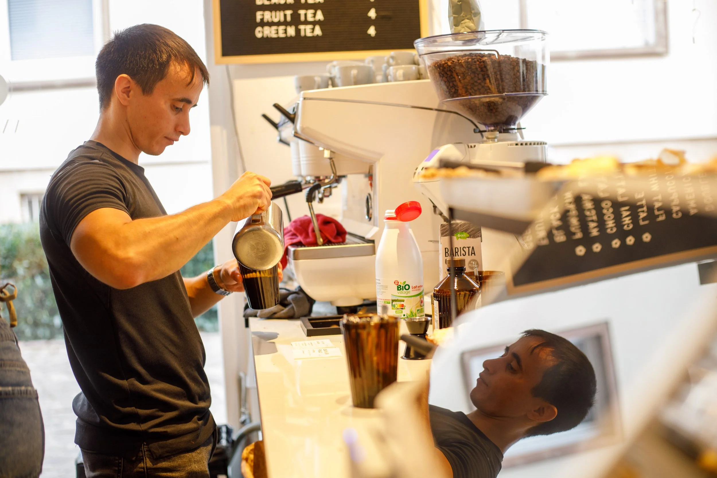 Un barista préparant un café derrière le comptoir d'une cafétéria, avec un jeune homme assis à la caisse en train de regarder sa reflection dans un miroir.