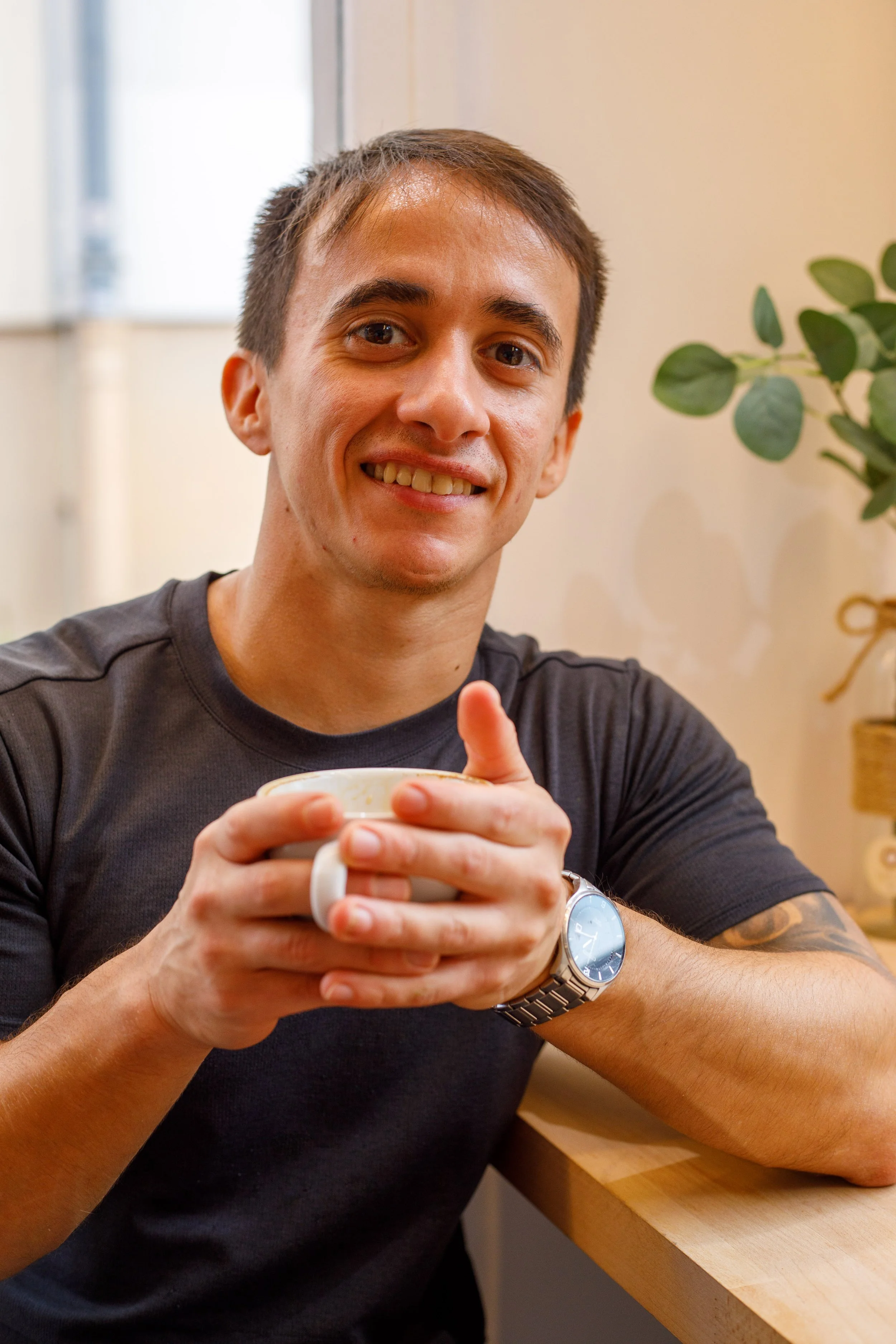 Homme souriant tenant une tasse à café, portant une montre en acier, assis à une table en bois, décor moderne avec plante verte en arrière-plan.