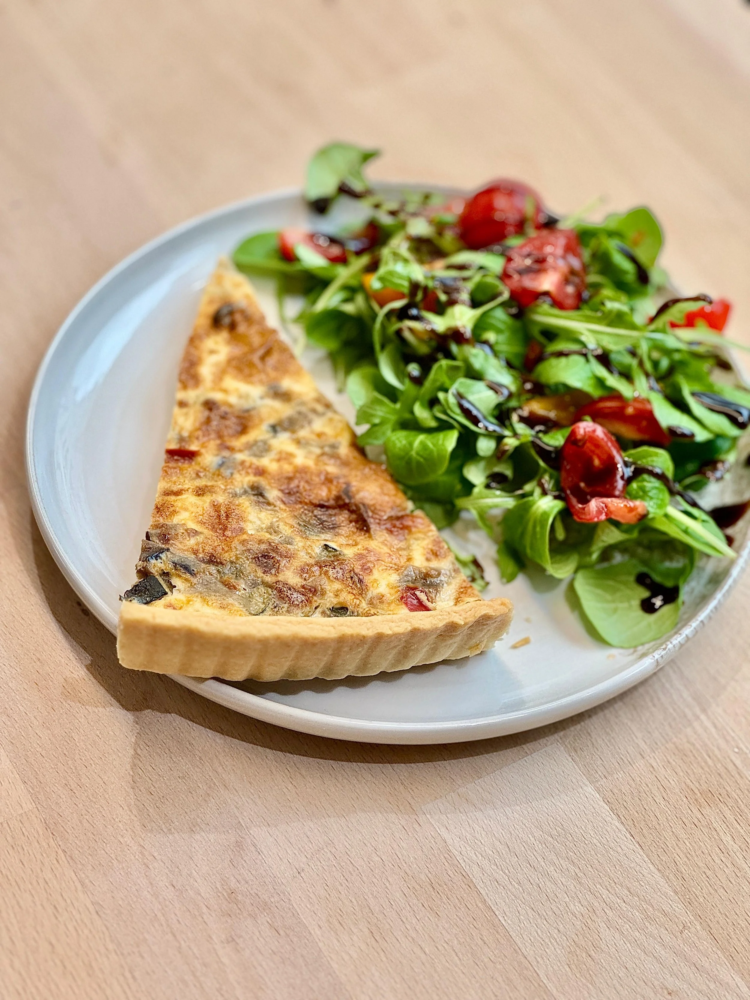 Une part de quiche aux légumes fait maison accompagnée d'une salade verte avec des tomates cerises et de la vinaigrette balsamique.