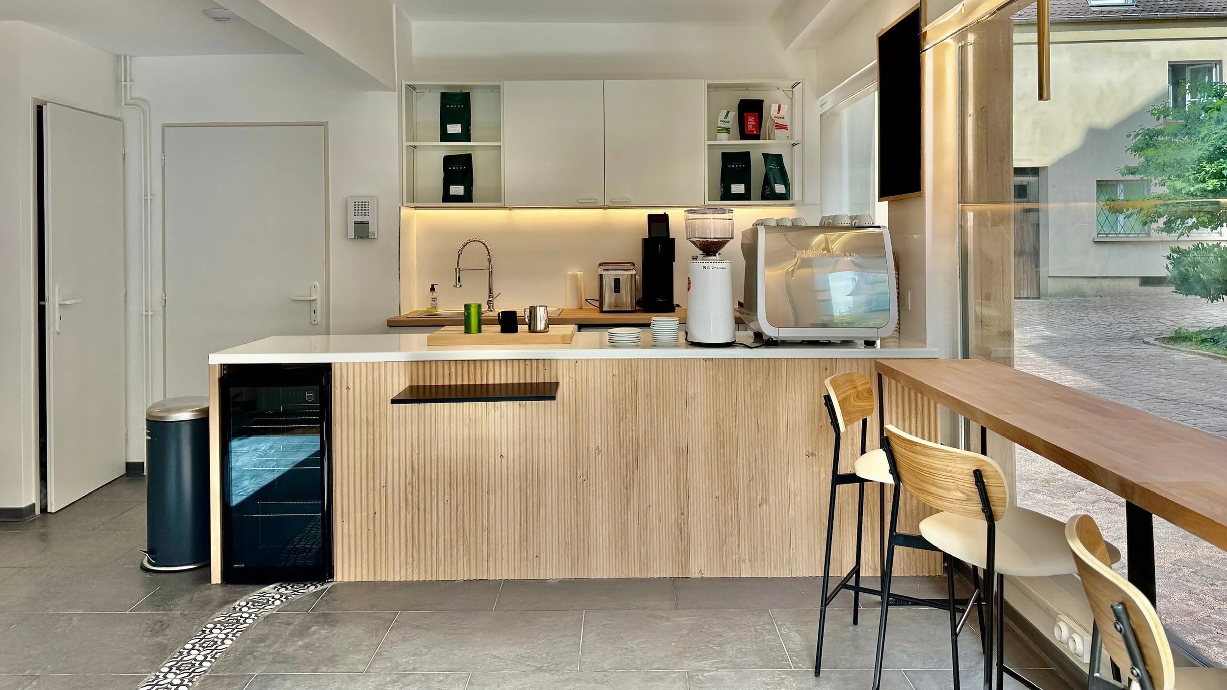 Cuisine moderne avec un comptoir en bois clair, un mini-réfrigérateur intégré, une machine à café, un blender et des tasses. Il y a aussi une grande fenêtre laissant entrer la lumière naturelle, avec des chaises de bar en bois et un comptoir en bois au bord de la fenêtre.