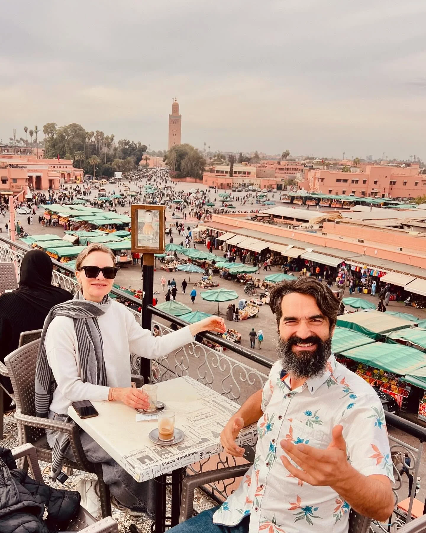 Deux touristes savourant une boisson à Marrakech avec la place Jemaa el-Fna, la mosquée Koutoubia en arrière-plan et le marché de Marrakech en bas.