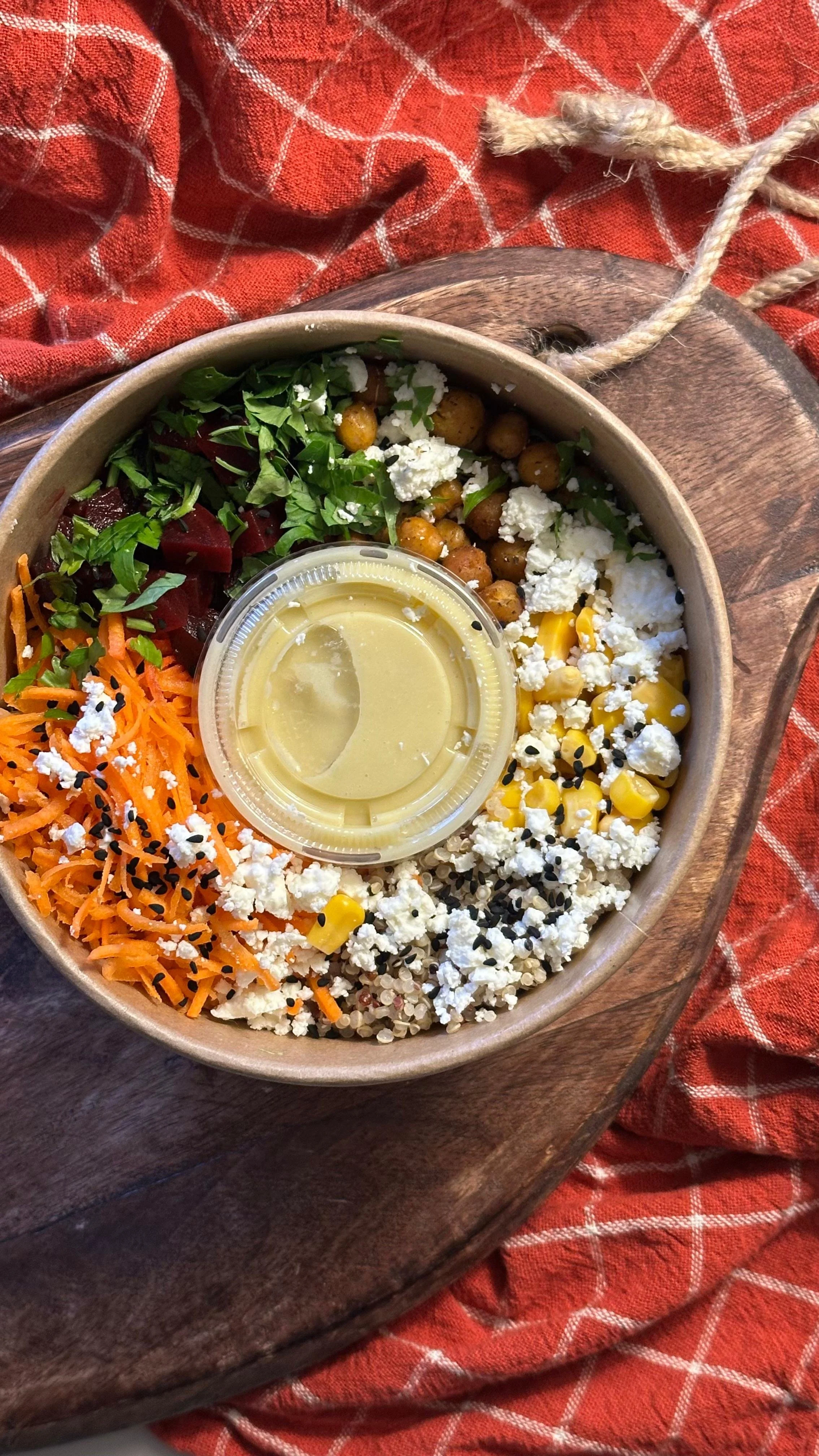 Bol de salade avec un base quinoa avec des légumes variés : carotte, betterave, mais, persil, fromage feta, graines, et une petite vinaigrette maison à la moutarde, placé sur une planche en bois avec une serviette à carreaux orange en arrière-plan.
