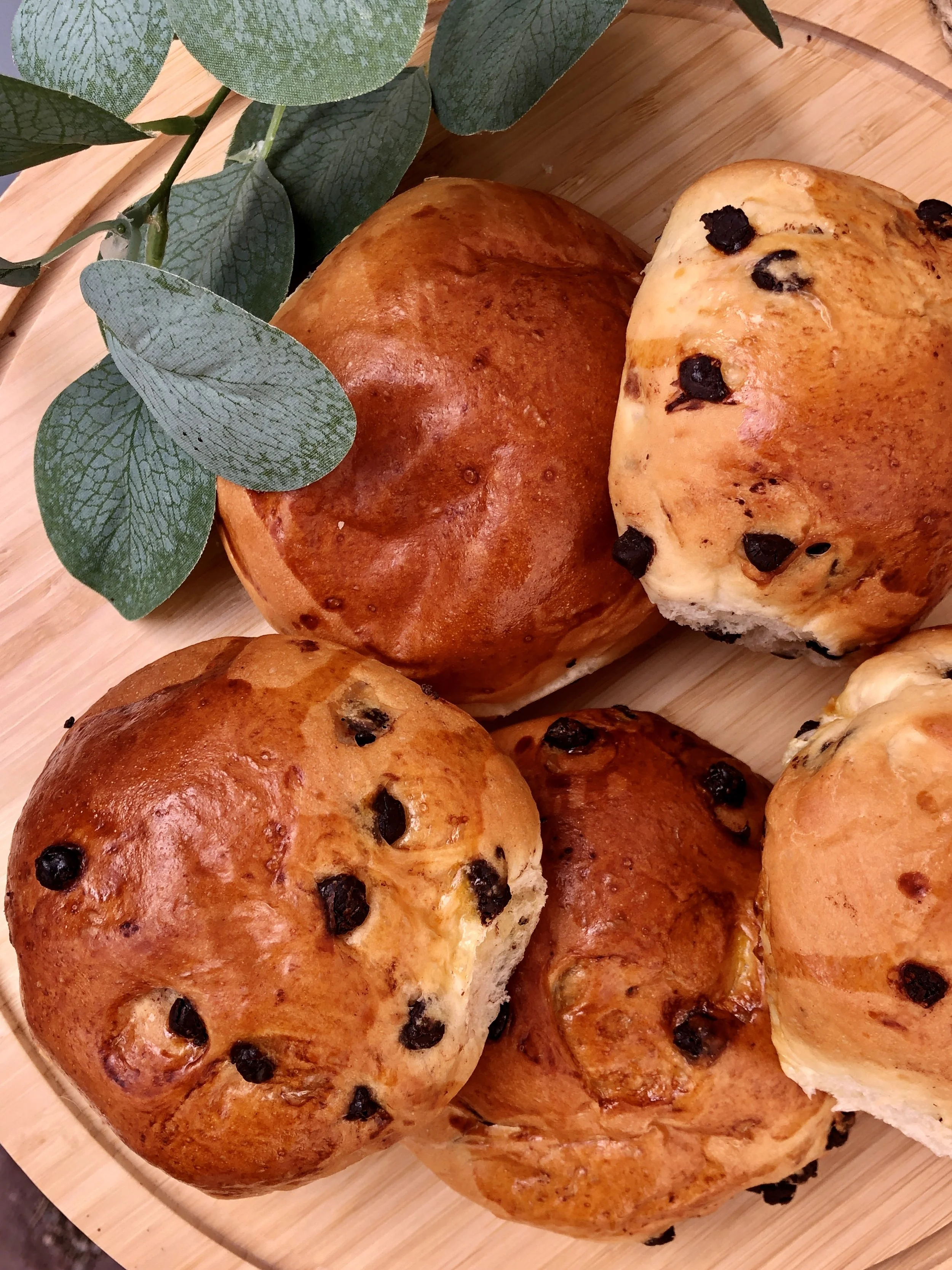 Petits pains briochés nature et aux pépites de chocolat sur une planche en bois avec des feuilles vertes en décoration.