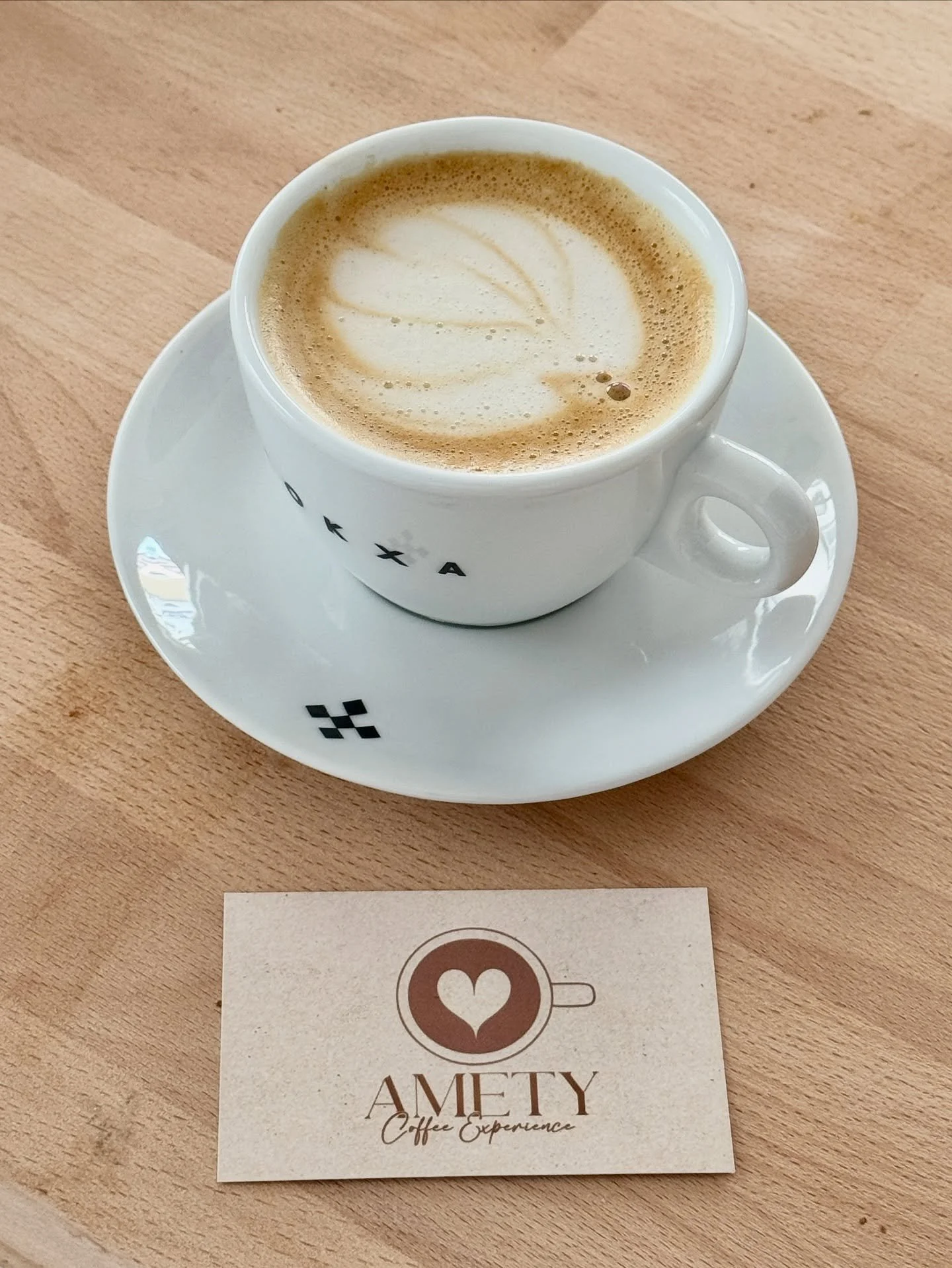 Une tasse de cappuccino avec un dessin de feuille en latte art sur la mousse, sur une table en bois clair, accompagnée d'une carte de visite avec le logo de tasse de café avec un cœur et le texte "AMETY Coffee Experience".