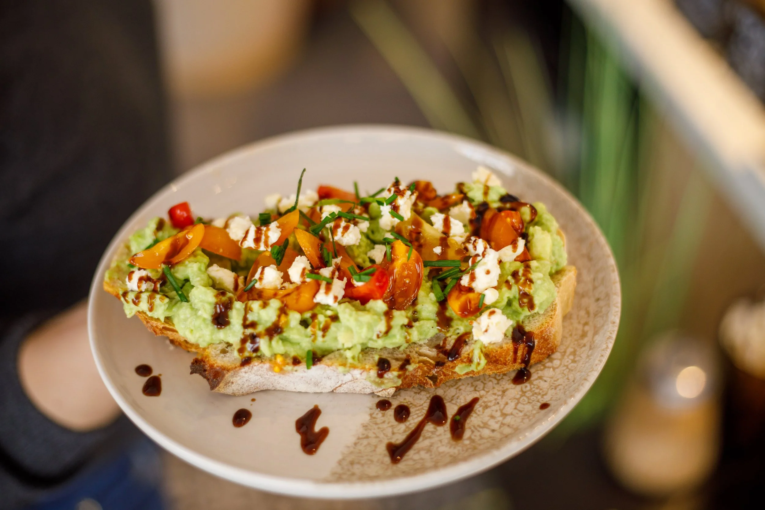 Tranche de pain garni d'avocat, tomates cerises, fromage feta, et sauce balsamique, sur une assiette blanche. Fait maison avec amour.