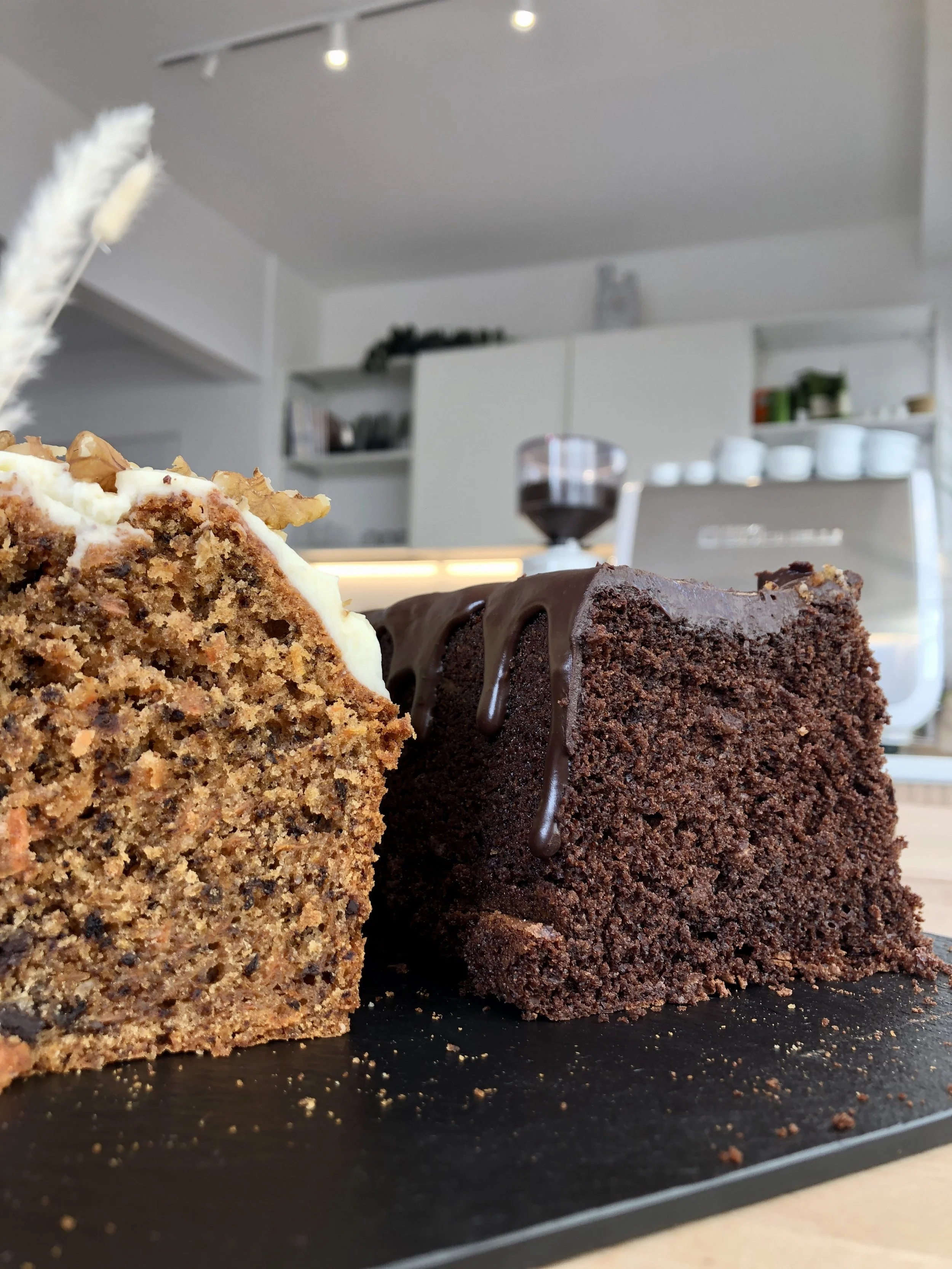 Deux cakes, un au chocolat avec un glaçage au chocolat, et un autre à la carotte avec glaçage crème cheese, sur une planche en ardoise, avec un fond de cuisine moderne de café.