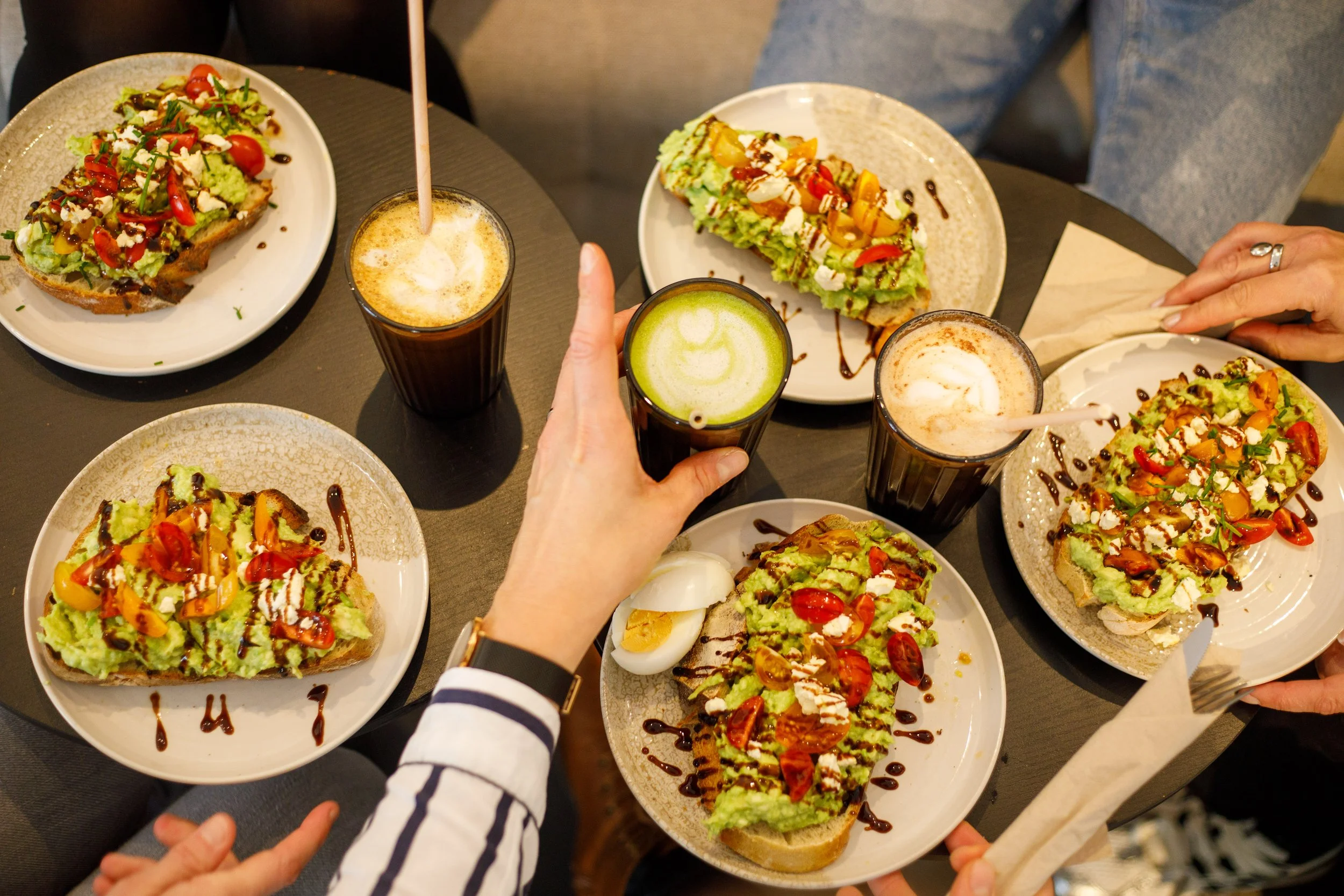 Avocado toast : tartines garnies d'avocat, tomates cerises, feta et de sauces balsamique, accompagnées de matcha latte et de tchai latte, sur une table du café à partager entre amis.