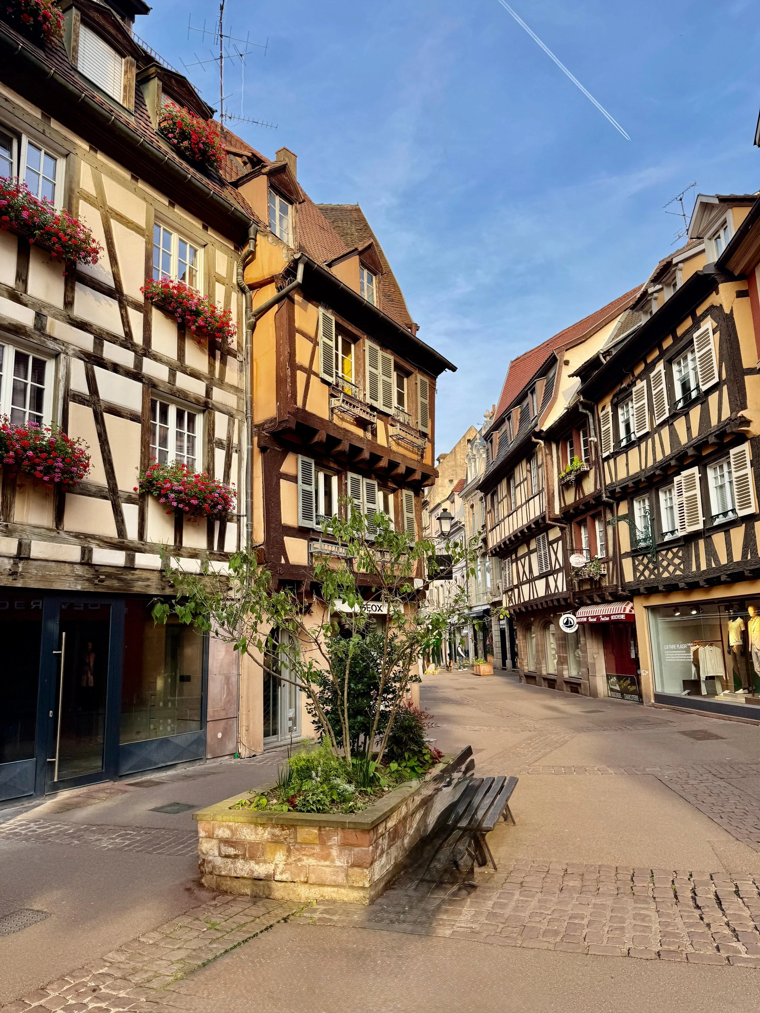 Rue pavée avec des bâtiments à colombages colorés, fleurs en balcon et un banc en bois devant un arbre dans une ville pittoresque.