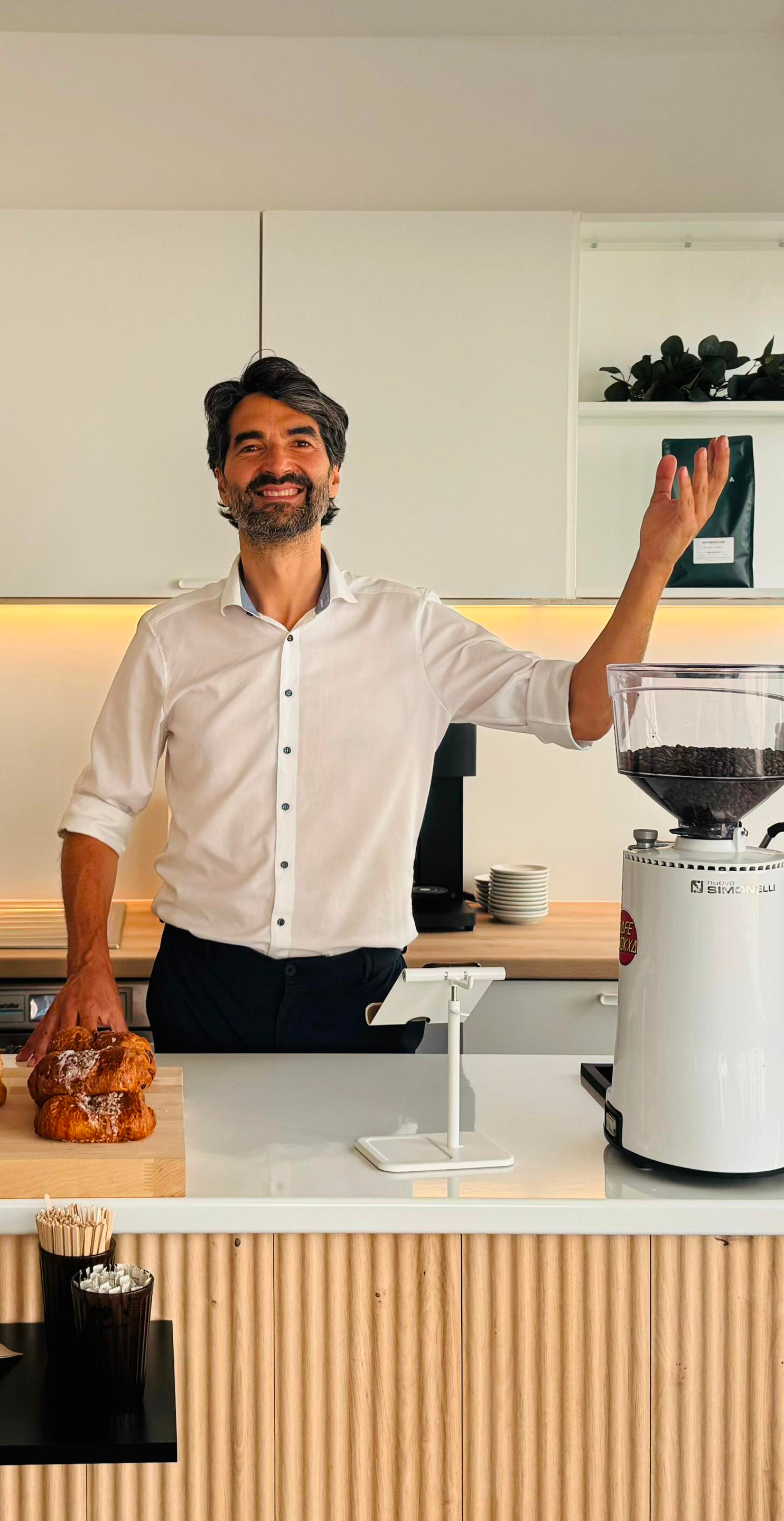 Un homme souriant en chemise blanche dans une cuisine, à côté d'une machine à café enroulée de grains de café, avec des croissants sur le comptoir et des tasses empilées en arrière-plan.