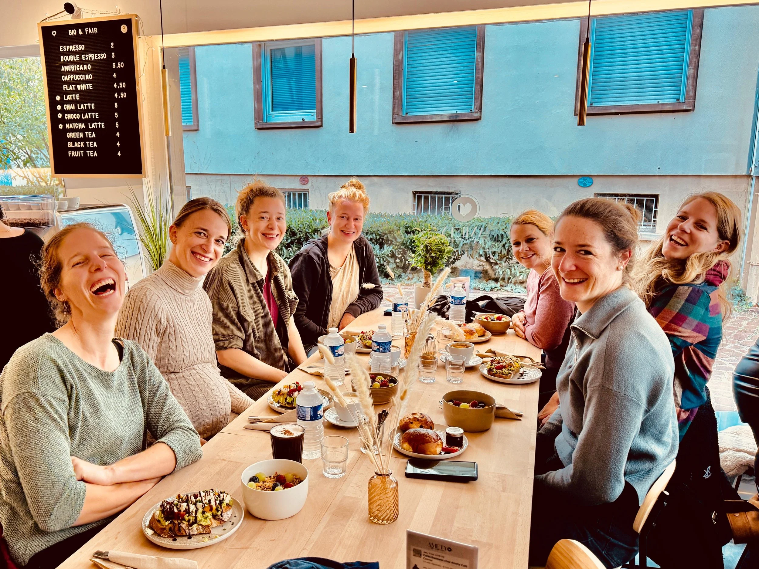 Groupe de huit femmes souriantes assises à une table dans un café, partageant un repas, avec un menu à l'arrière-plan et des fenêtres donnant sur une façade urbaine.