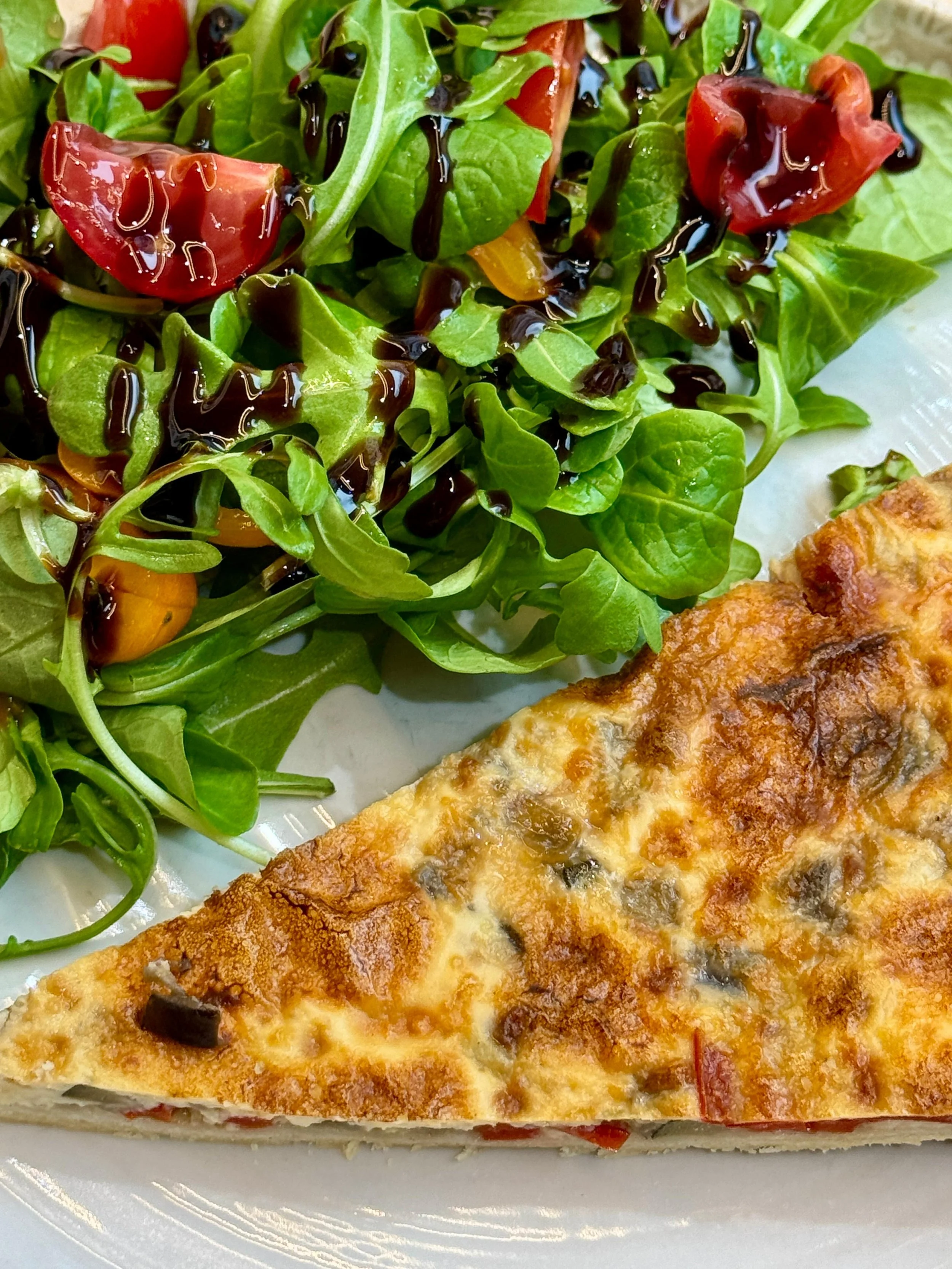 Une tranche de quiche aux légumes accompagnée d'une salade verte avec des tomates cerises et de la vinaigrette balsamique.
