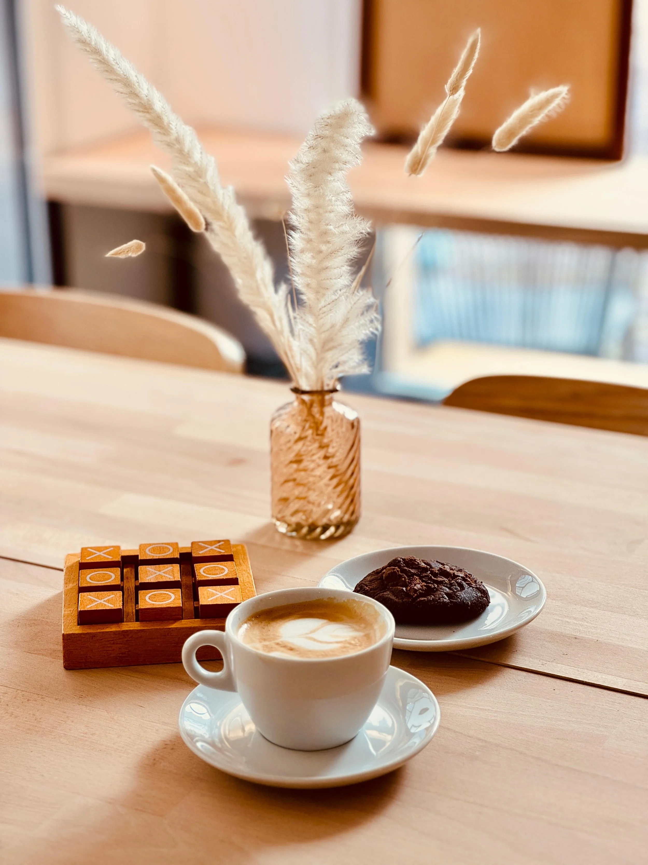 Une tasse de café cappuccino, un cookie chocolat et pépites de chocolats sur une petite assiette, un jeu de morpion en bois, et un vase avec des herbes de décoration, sur une table en bois.