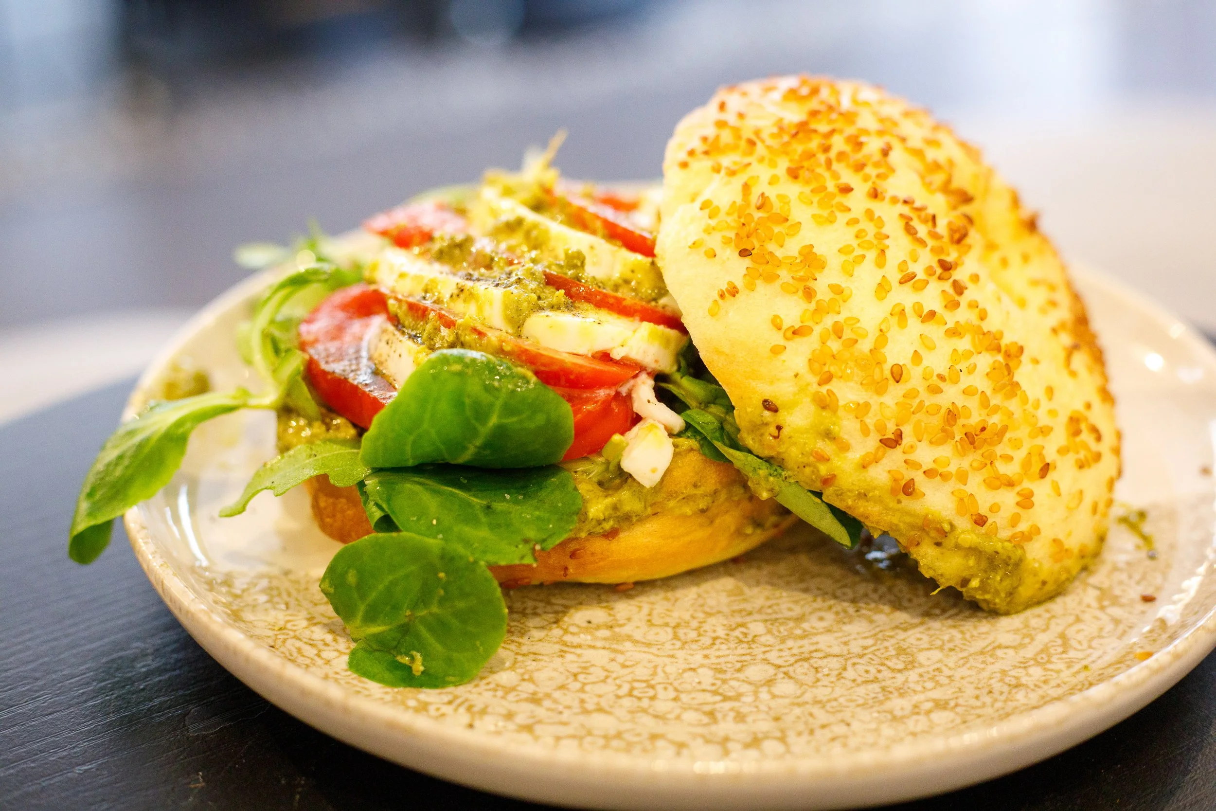 Sandwich végétarien avec pain bagel, tomates, mozzarella, salade et sauce pesto, servi sur une assiette.