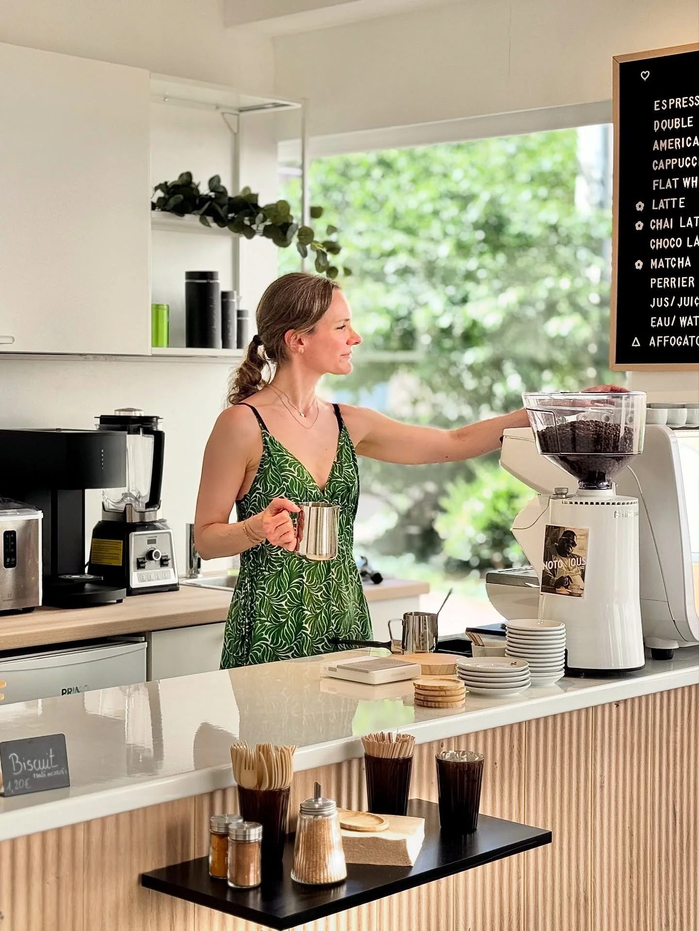 Femme au comptoir de café dans un établissement lumineux, portant une robe verte avec un motif de feuilles, préparant une boisson derrière un bar moderne.
