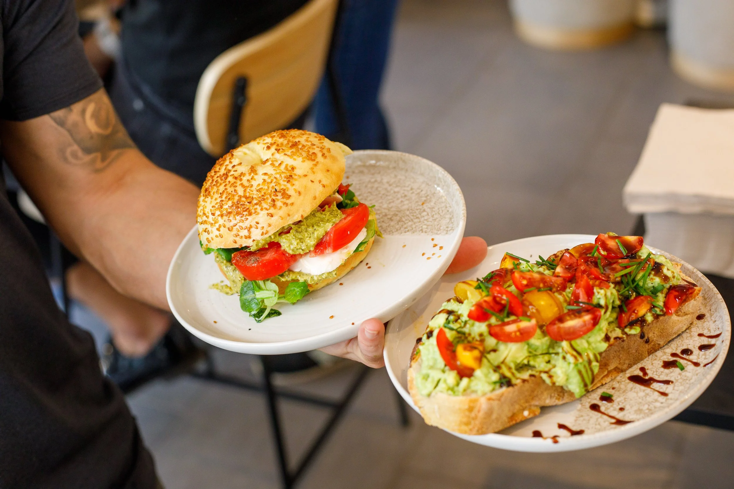 Une personne tient deux assiettes de nourriture, une avec un pain bagel aux graines avec de la laitue, de la tomate, de la mozzarella et de la sauce pesto, et une autre avec un avocado toast garnie de guacamole de tomates cerises de sauce balsamique.