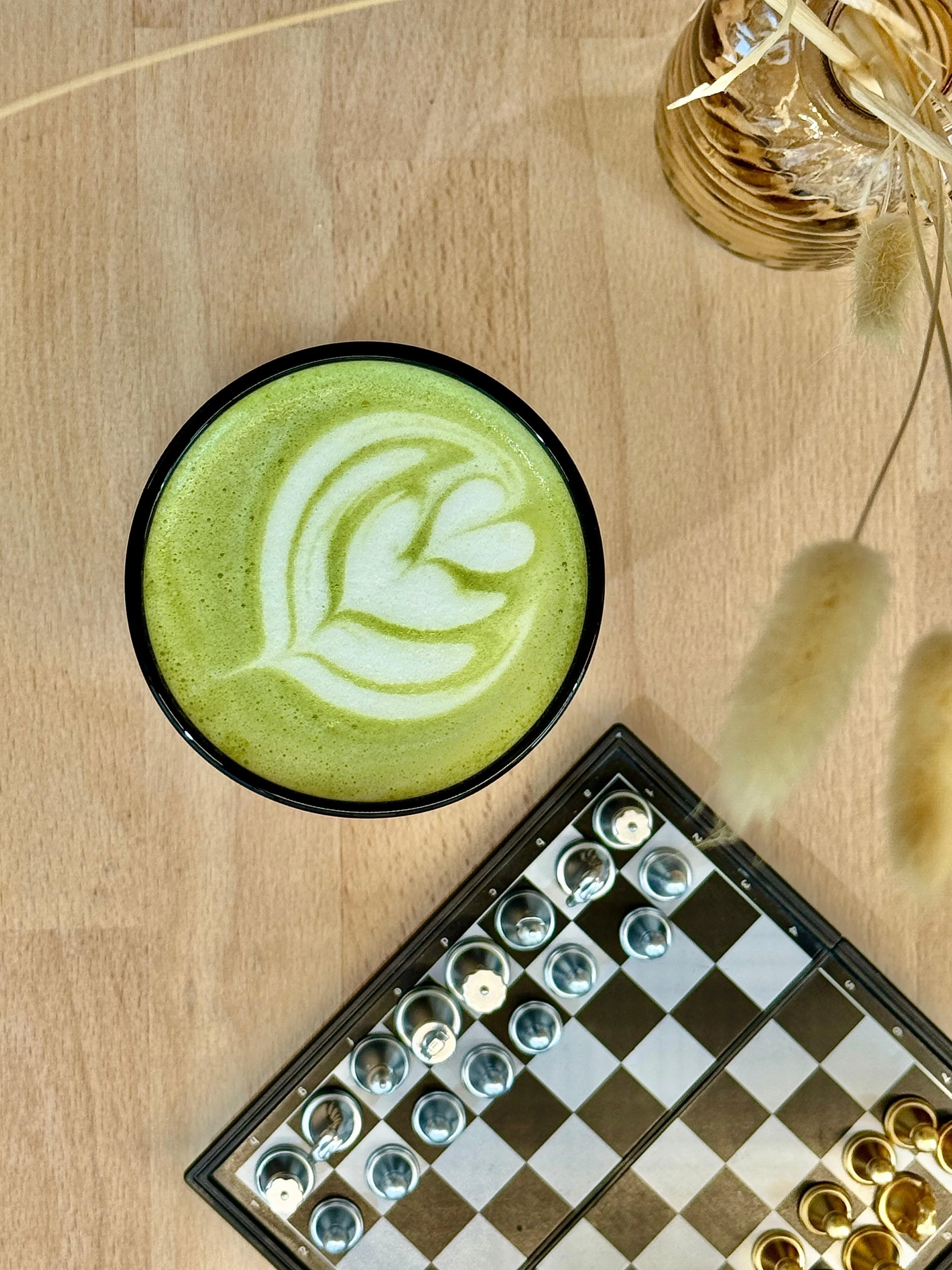 Verre de matcha latte avec latte art en forme de plusieurs cœurs les uns dans les autres, jeu de société échec, décor floral en finition sur une table en bois.