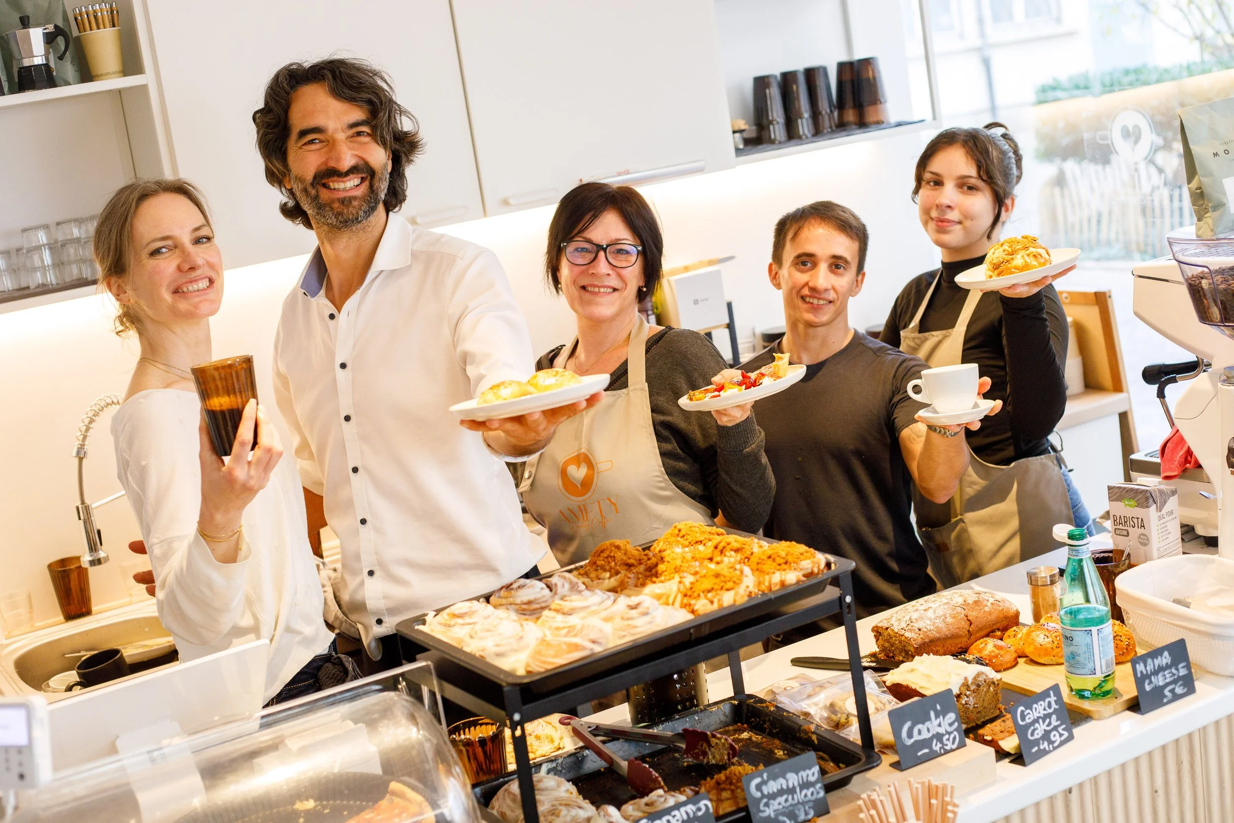 Groupe de cinq personnes dans une ambiance chaleureuse de café ou pâtisserie, posant derrière un comptoir avec diverses pâtisseries et cafés, tous souriant et tenant des assiettes ou des tasses.