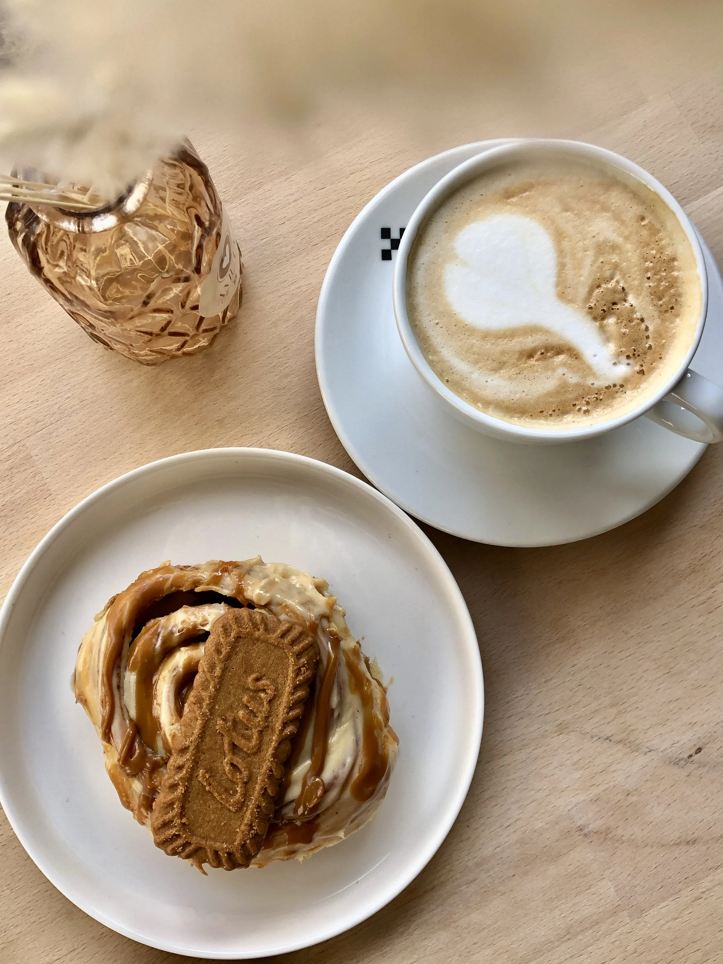 Une tasse de café avec de la mousse, un dessert avec un biscuit et un caramel, et une bouteille en verre oxydé sur une table en bois.